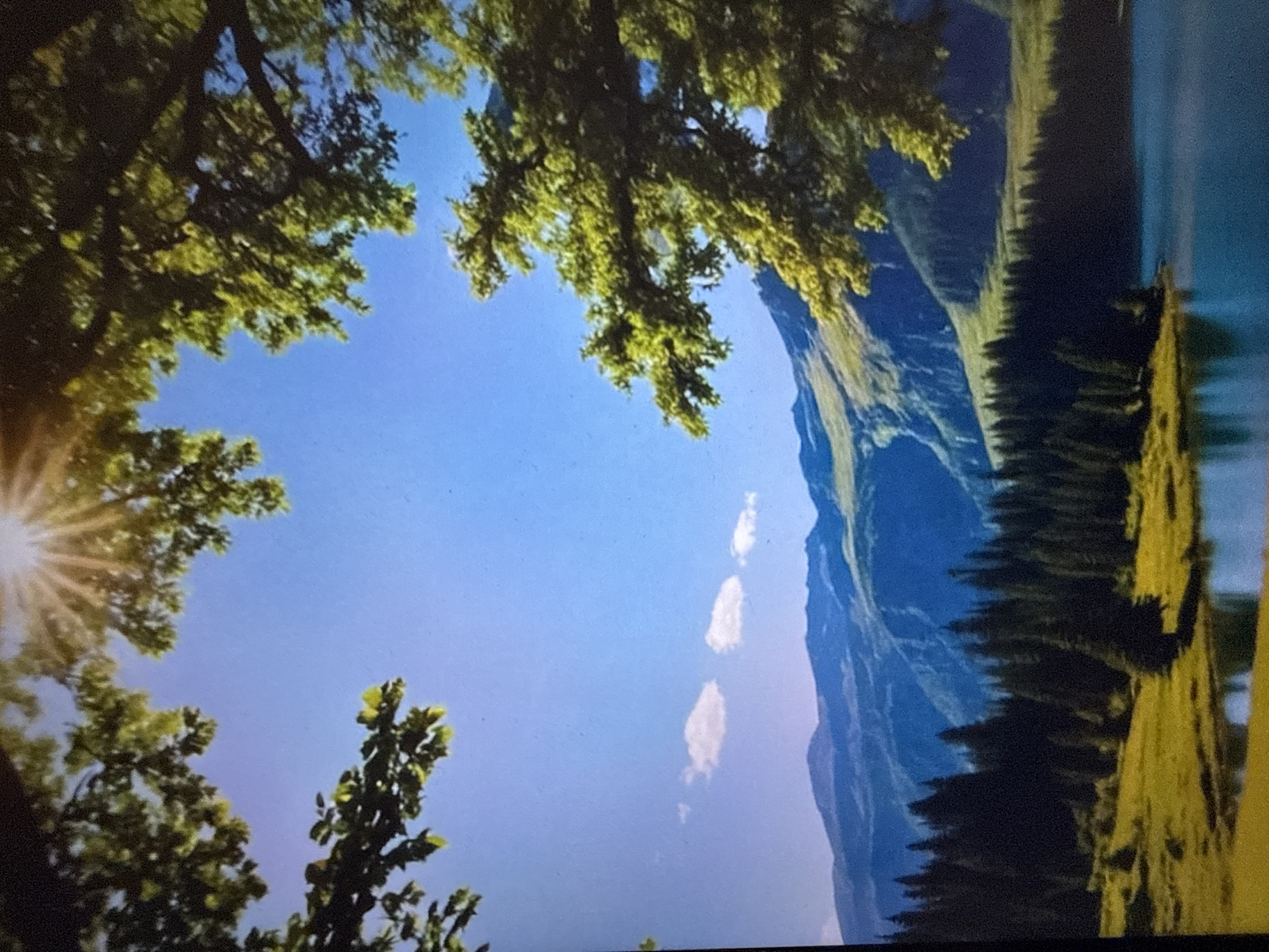 This image depicts a serene mountain landscape. In the foreground, lush green trees frame the scene from the top and right. Beyond the trees, a clear blue sky is visible, with the sun's rays streaming down from the upper left corner. A few scattered white clouds drift across the sky. In the middle ground, a body of water reflects the sky and the surrounding landscape. Beyond the water, a dense forest of dark green pine trees stretches across the terrain. Further in the background, rolling green hills rise up, leading to majestic blue mountains that recede into the distance, their peaks shrouded in a soft haze. The overall impression is one of natural beauty and tranquility.