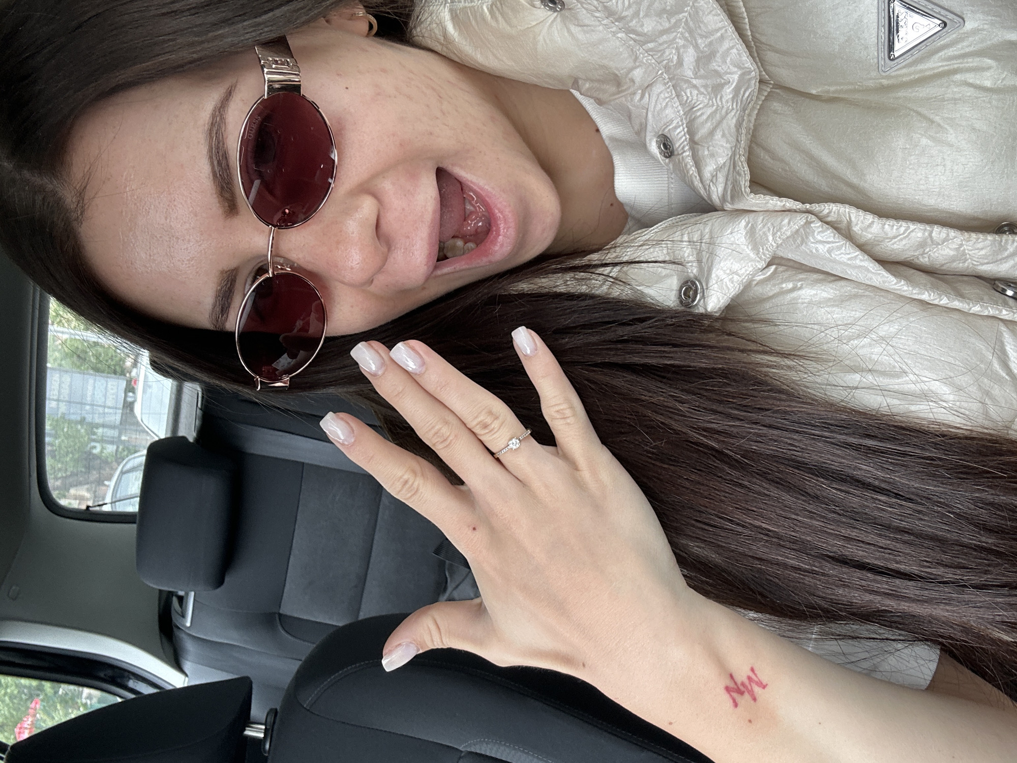 A young woman is sitting in a car, wearing a white puffer jacket and sunglasses. She has long, dark hair and manicured fingernails, and she is wearing a ring on her left ring finger. She has a tattoo on her arm. The car's interior is visible, showing black seats and a window. The location appears to be within a vehicle, there is no clear indication that this is within the city of Plovdiv, Bulgaria.