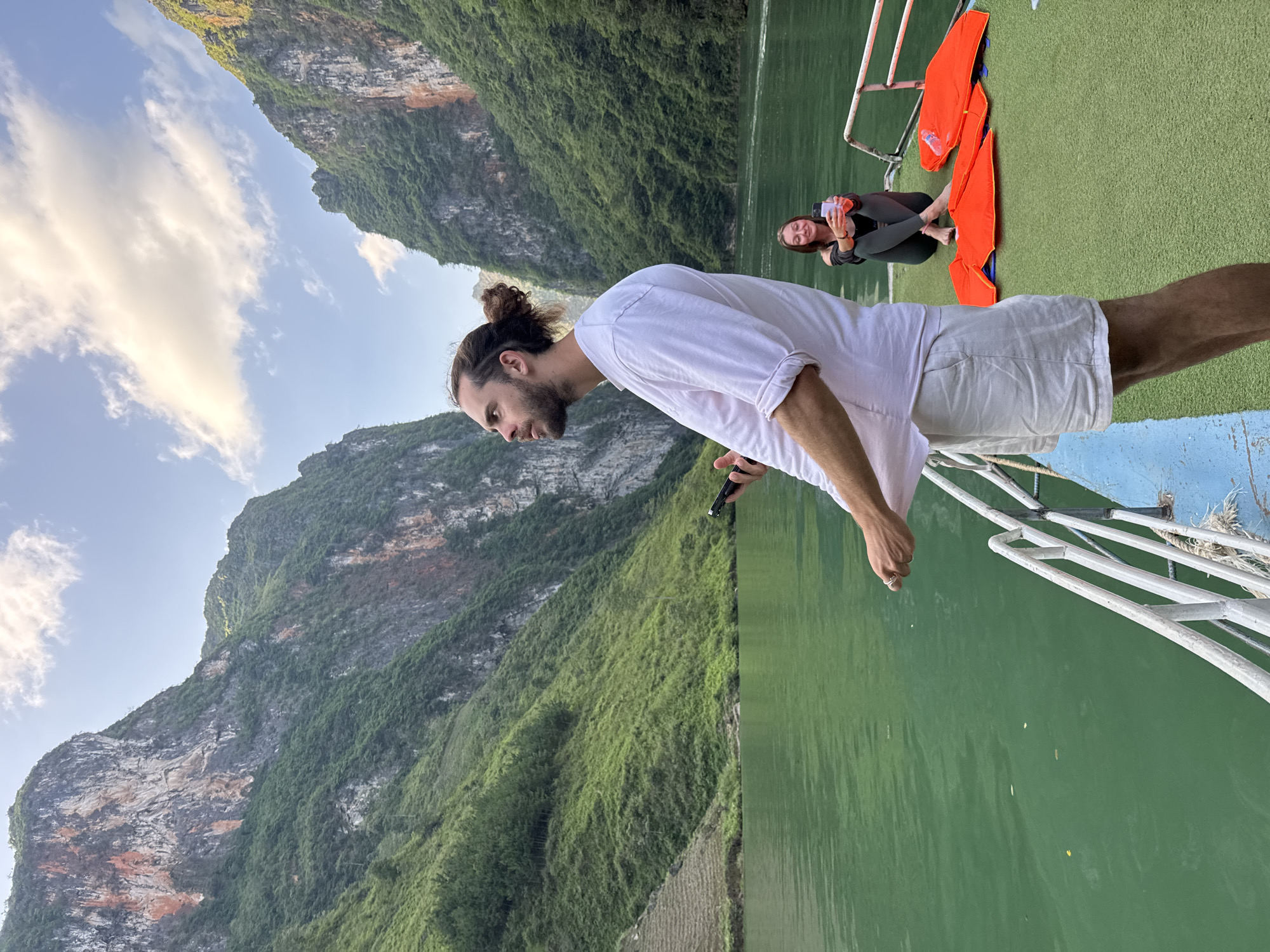 The image depicts a man with long hair tied in a bun, wearing a white t-shirt and light shorts, leaning over the railing of a boat on a body of water surrounded by lush, green mountains. The water appears calm and green. Another person, a woman, is sitting cross-legged further back on the boat, looking at a phone or camera. Orange life jackets are piled nearby. The backdrop features rocky mountains covered in dense vegetation and a partly cloudy sky. The location is likely in or near the city of Đồng Văn Commune, Vietnam, given the landscape features.