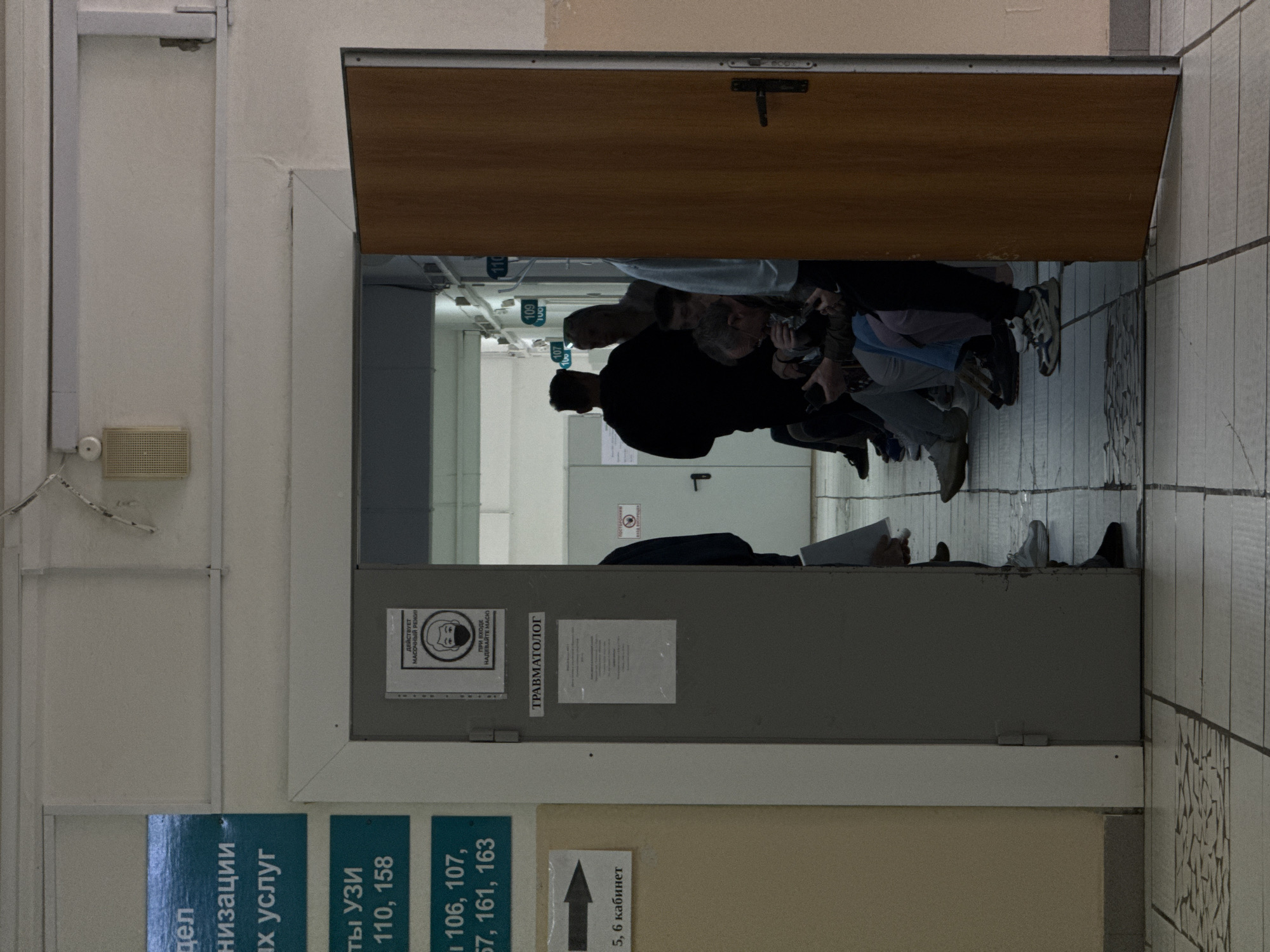 The image, taken in Chaikovsky, Russia, captures a view through an open wooden door into a bustling waiting area, likely a medical clinic or hospital corridor. The perspective is from just outside the room, looking inwards, with the door acting as a visual frame.
Several people are visible inside, seated along a wall, suggesting they are waiting for appointments. Their faces are partially obscured or seen in profile, and they appear calm, some looking forward, others down. The setting is brightly lit by interior fluorescent lighting, typical of a public building. The floor in the foreground is tiled in a light pattern, transitioning to a different, possibly worn, floor surface inside the waiting room.
On the wall to the left of the door, there's a light-colored speaker or intercom box and several informational signs. These blue signs with white Russian text indicate departments or room numbers, such as "Ultrasound rooms 110, 158" and a list of other room numbers like "106, 107, 57, 161, 163". An arrow sign points towards "5, 6 кабинет" (rooms 5, 6).
Attached to the grey doorframe, another sign displays a mask icon with text in Russian: "ДЕЙСТВУЕТ МАСОЧНЫЙ РЕЖИМ" (MASK REGIME IS IN EFFECT) and "ПРИ ВХОДЕ НАДЕВАЙТЕ МАСК" (WHEN ENTERING WEAR A MASK), indicating health precautions. Below this, the word "ТРАВМАТОЛОГ" (TRAUMATOLOGIST) is prominently displayed, identifying the type of medical service available in this section. Further inside, numbers "110", "109", and "107" are visible on the walls, likely marking different consultation rooms. The overall atmosphere is one of routine waiting in a functional medical environment.