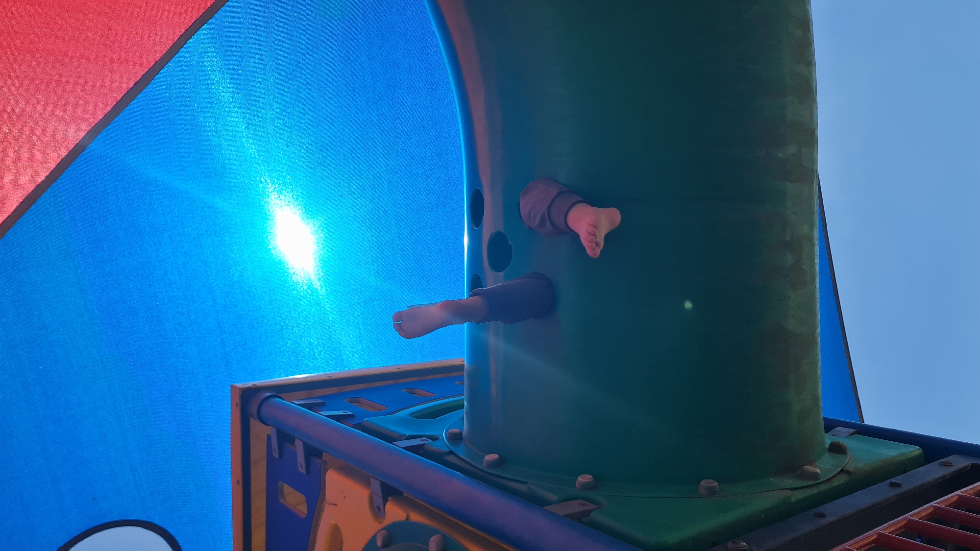 A low-angle shot of a playground structure with red, blue, and green elements. Two children's bare feet protrude from round openings in a green cylinder, which sits on top of a platform with blue and yellow elements. The background includes a blue sky and a bright light source reflected in the blue fabric of the playground structure. The location is Lachish Regional Council, Israel.