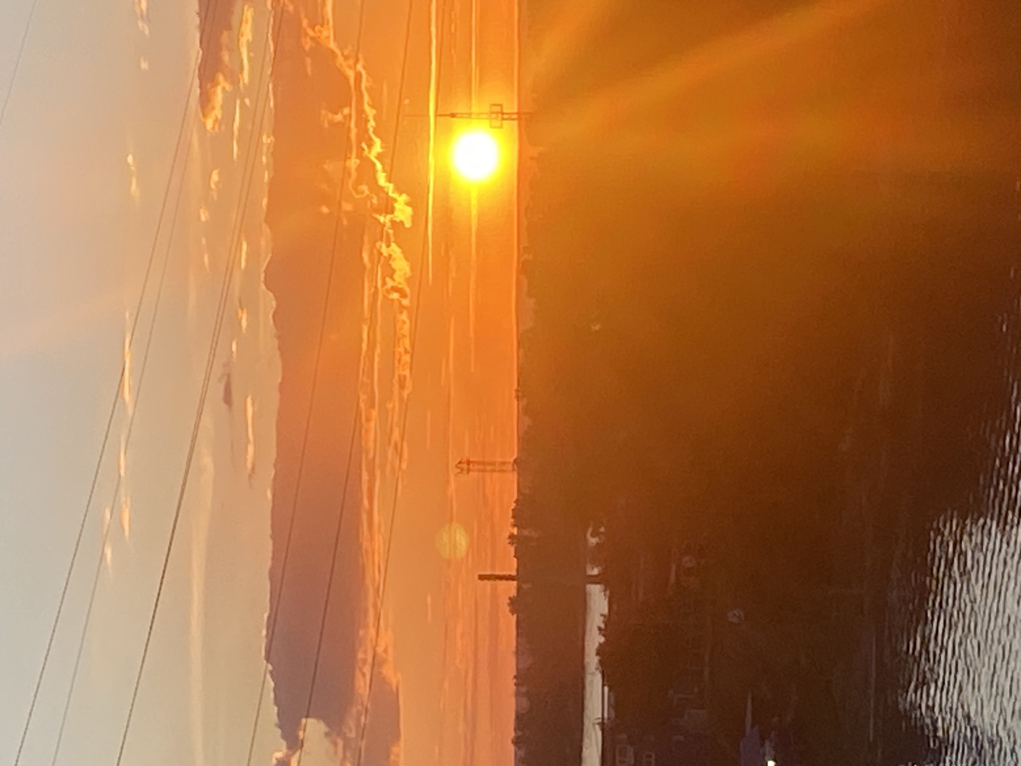 The image depicts a sunset scene in Novokuybyshevsk, Russia. The sun, a bright, glowing orb, is setting over a body of water, casting a warm, orange and yellow light across the sky and reflecting on the rippling surface of the water. The water is dark, contrasting with the vibrant sky.
On the left side of the image, there are power lines stretching across the frame, with tall, industrial structures visible in the background, silhouetted against the bright sky. These structures suggest an industrial or urban setting. The sky itself is filled with wispy clouds that are illuminated by the setting sun, adding texture and depth to the scene.
The overall mood of the image is serene and picturesque, capturing the beauty of a sunset in an industrial landscape. The time of day is clearly sunset, indicated by the low position of the sun and the warm, saturated colors in the sky. There is no visible text in the image.