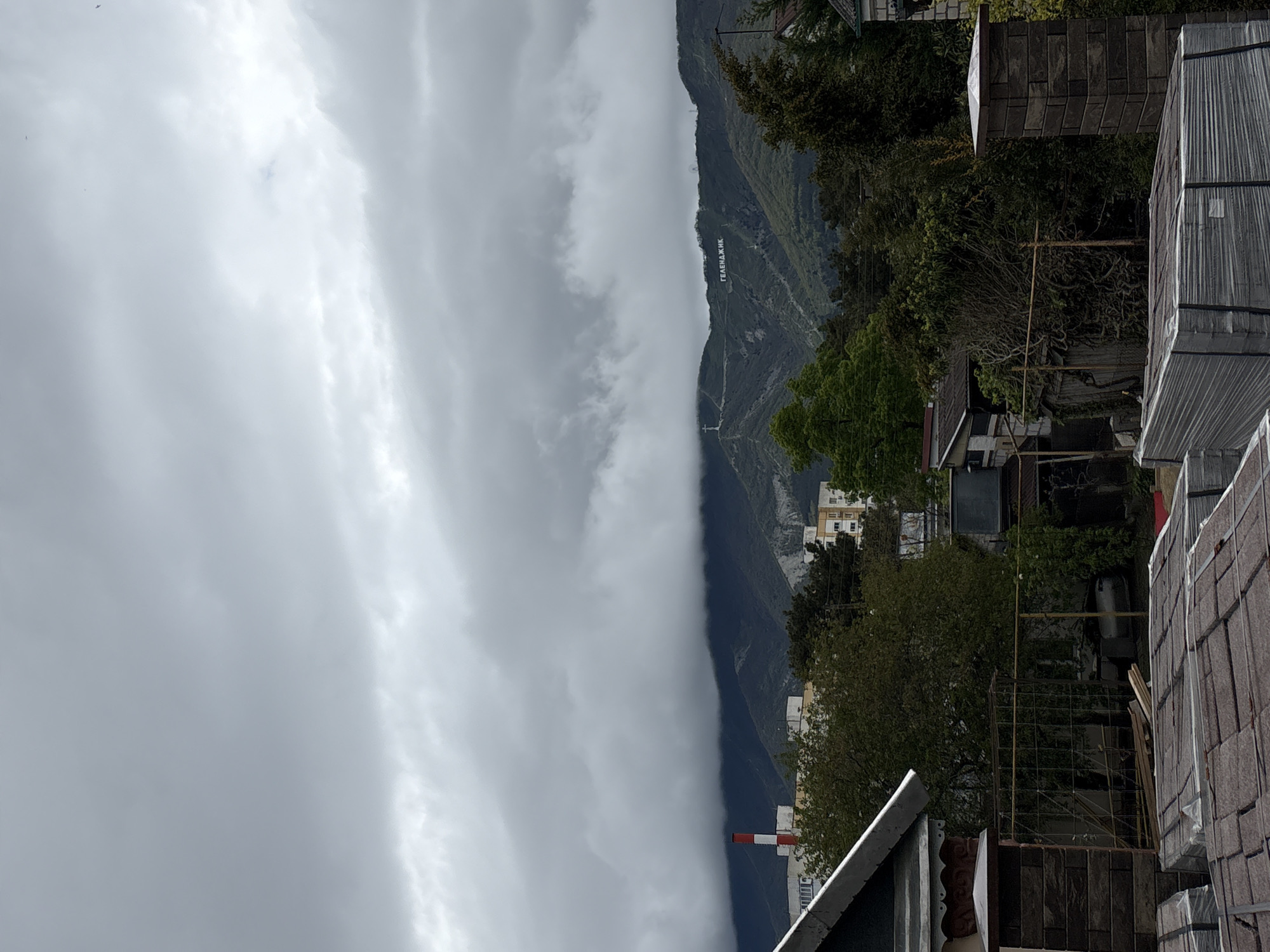 The image captures a cloudy day in Gelendzhik, Russia, with a view of the mountains. The word "ГЕЛЕНДЖИК" (Gelendzhik) is written in large white letters on the mountainside. Dense cloud cover is present, partially obscuring the view. The foreground includes buildings, trees, and construction materials, suggesting an urban or residential area at the foot of the mountain. A red and white striped smokestack is visible. The overall scene suggests a landscape where urban development meets mountainous terrain.