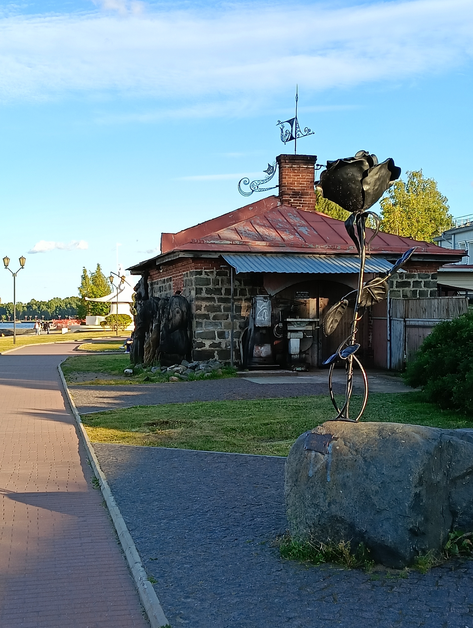 This image depicts a sculpture of a black metal rose on a large rock in front of a stone building with a red metal roof. A weather vane adorns the chimney of the building. To the left of the building, a paved walkway runs alongside a grassy area. In the background, a body of water with trees on the opposite shore can be seen, along with lampposts and people walking. The sky is blue with scattered clouds. This scene is located in Petrozavodsk, Russia, specifically on the Onega embankment.