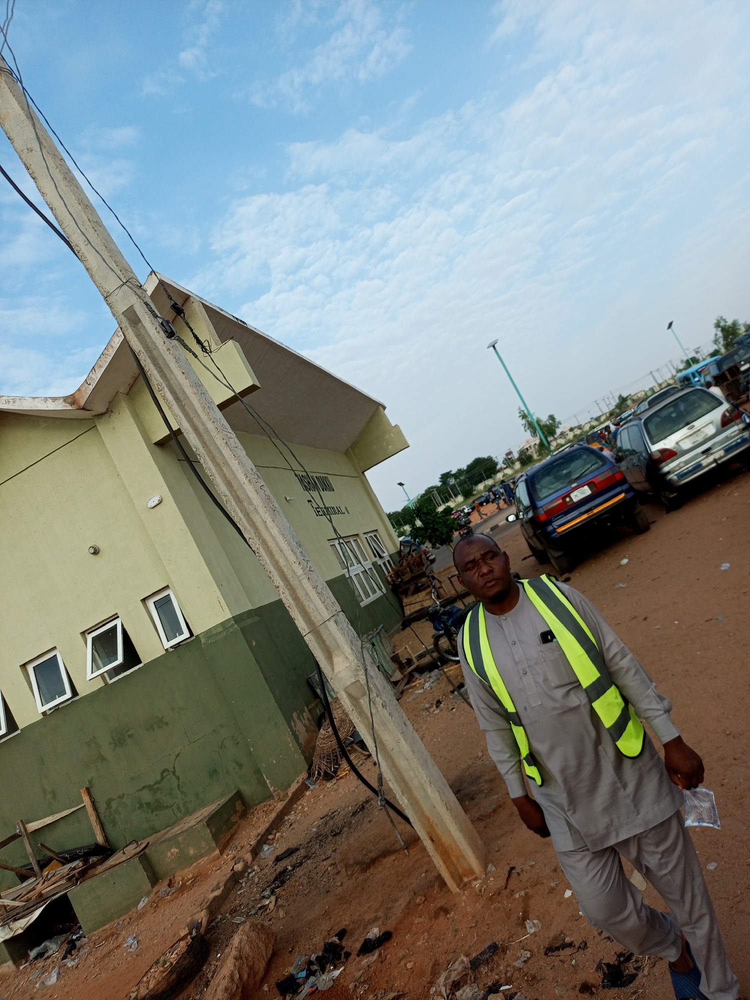 In Gombe, Nigeria, a man wearing a grey traditional outfit and a bright yellow-green safety vest stands on a dusty, unpaved lot beside a light green and dark green building. The building has multiple windows, some open. A concrete utility pole leans precariously to the left of the frame, wires strung from it. Beyond the man and the building, a parking lot is visible with several cars and a distant bus. The sky is a pale blue with scattered white clouds. The ground is littered with debris, including plastic bags and discarded items.