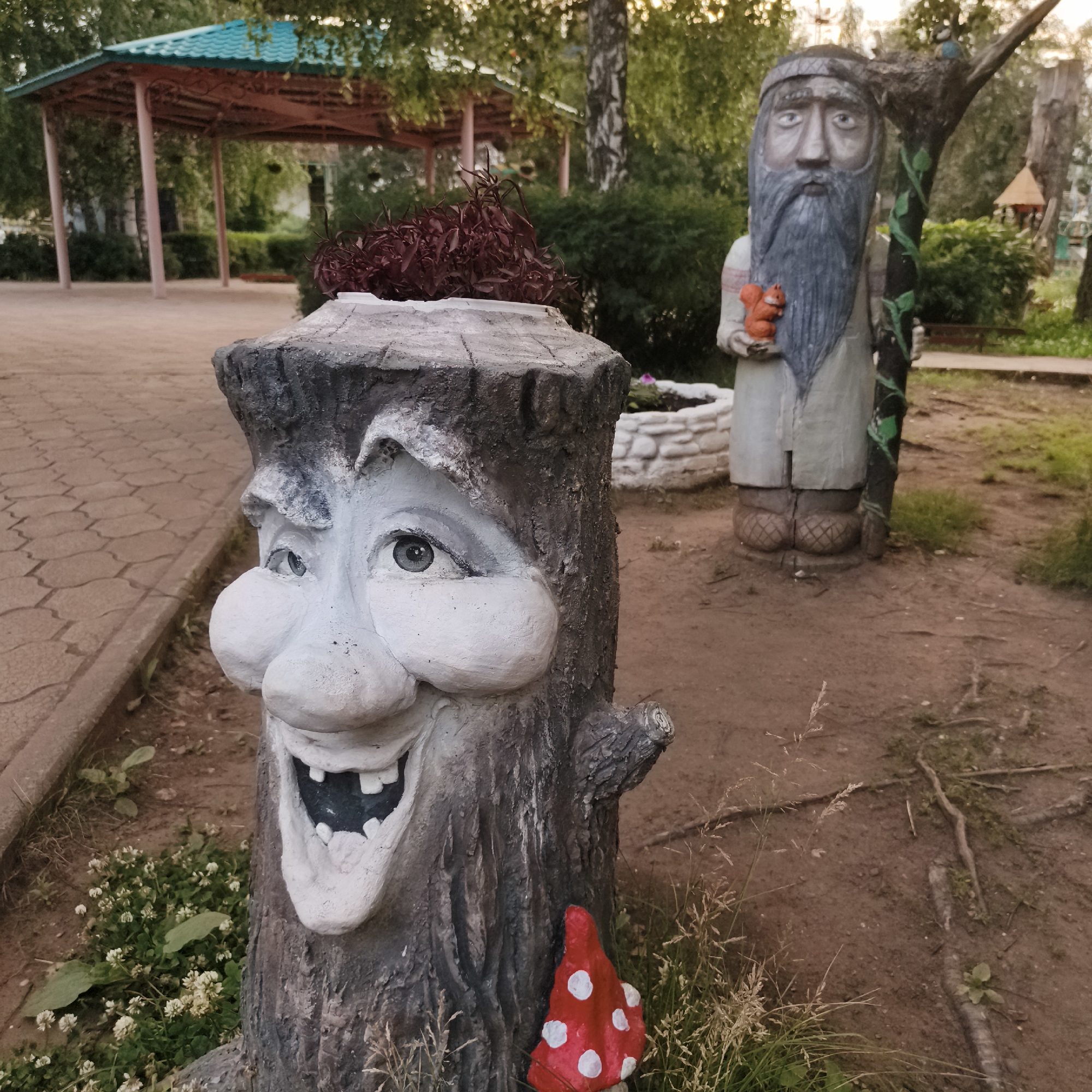 The image depicts a park in the city of Chaikovsky, Russia. In the foreground, a tree stump has been carved and painted to resemble a character with a large nose, eyes, and a wide, toothy grin. It has a planter on top filled with reddish foliage. To the right, further back, is a wooden statue of a bearded man in a long robe, holding a small orange squirrel. He has a band around his forehead and is standing next to a tree trunk with decorative green vines. Behind these figures, a turquoise-roofed gazebo with ornate metal supports is visible, surrounded by green bushes and trees. The ground is a mix of dirt and paved walkway. A small red mushroom decoration with white spots is placed at the base of the tree stump character.