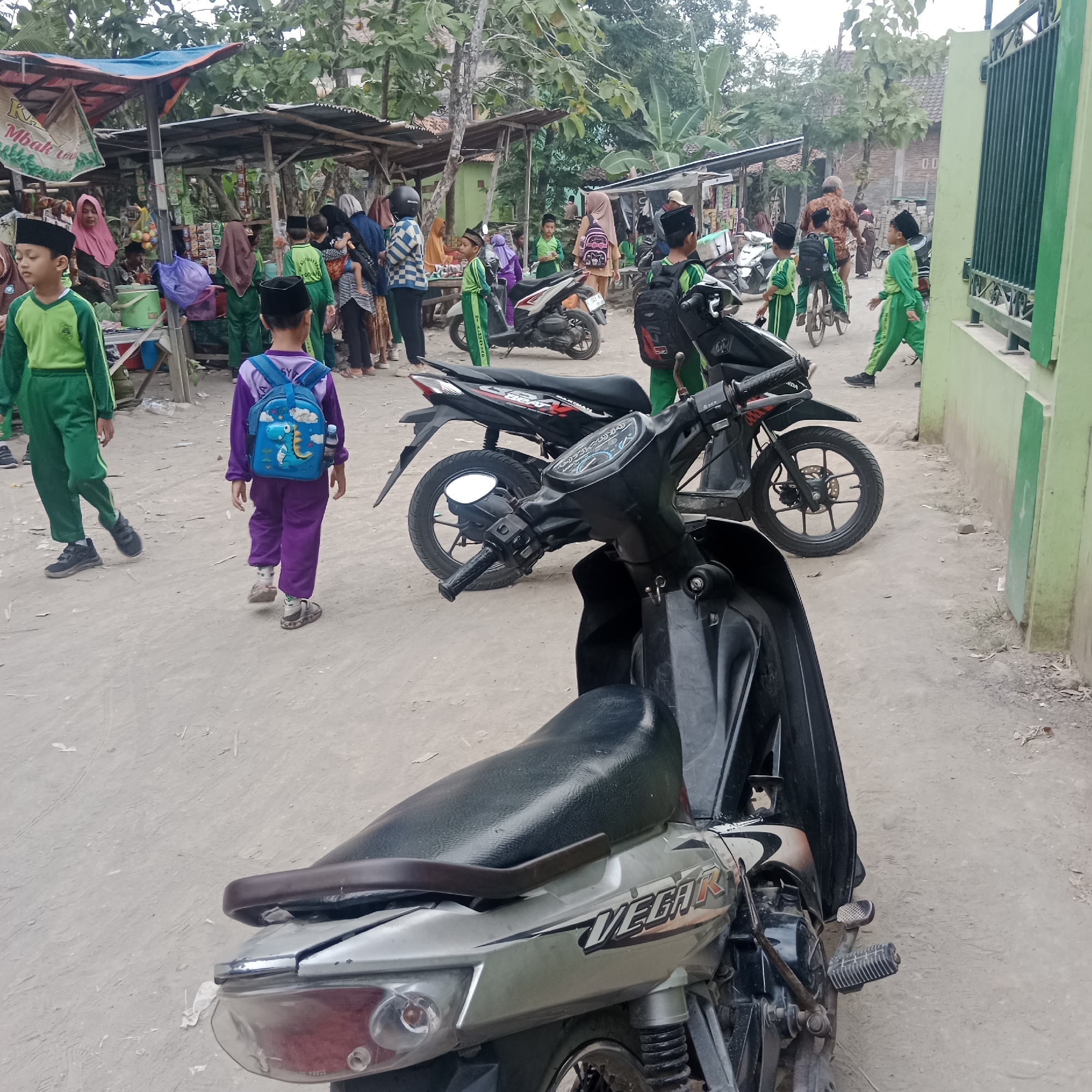 In the city of Tegowanu Kulon, Indonesia, a street scene unfolds with a group of children, some wearing green school uniforms and black peci hats, and others in purple attire. A young boy in a purple uniform carries a blue backpack adorned with a dinosaur design. Several motorcycles are parked on a dusty, unpaved road. Behind the children, makeshift stalls with awnings are set up, displaying various goods. The stalls are constructed with wood and fabric. In the background, trees and some buildings are visible, suggesting a semi-rural or suburban environment. The overall atmosphere appears to be one of daily activity, possibly after school, with the children milling about near the stalls and motorcycles.