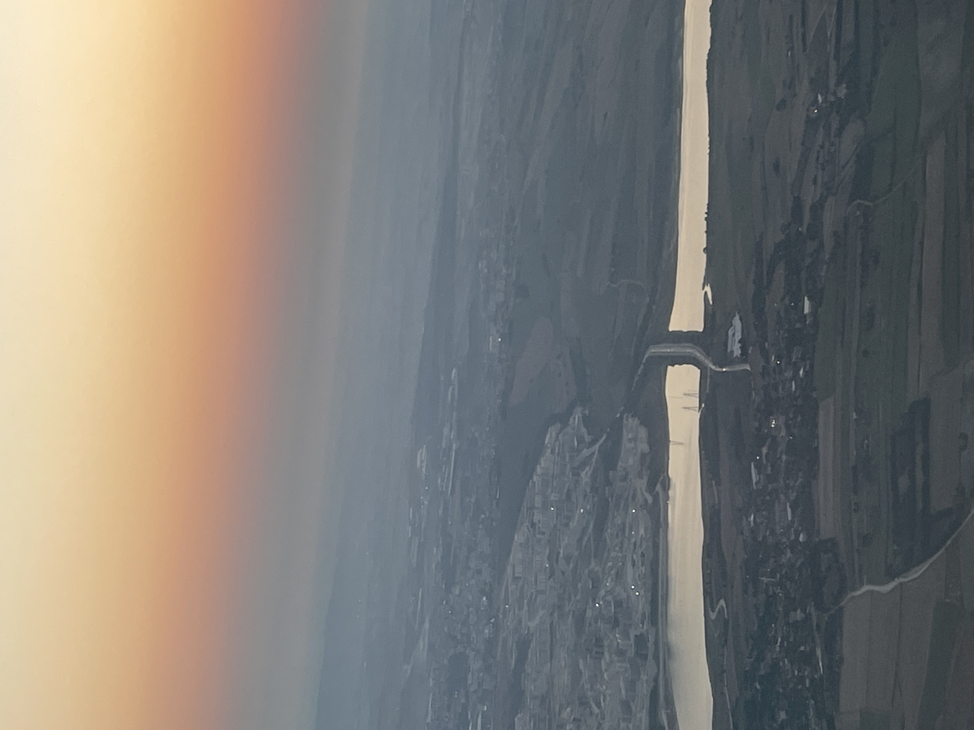 This aerial photograph captures a landscape during a sunset or sunrise, with the sky transitioning from a pale orange hue at the top to a hazy gray towards the horizon. The scene depicts a body of water, likely a reservoir or a lake, reflecting the sky's colors. A bridge spans across a section of this water. To the left of the water, a densely populated urban area with numerous buildings is visible, characterized by a grid-like layout. Beyond the urban sprawl, more distant hills and a hazy atmosphere obscure finer details of the landscape. To the right of the water, the terrain is less developed, showing agricultural fields and a scattering of houses or smaller settlements. The overall impression is one of an urban area adjacent to a natural water feature, with a sprawling landscape extending into the distance under a colorful, atmospheric sky. This scene is consistent with the geography of Beykoz, Turkey, which is known for its position on the Bosphorus Strait and its blend of urban development, natural landscapes, and water bodies.