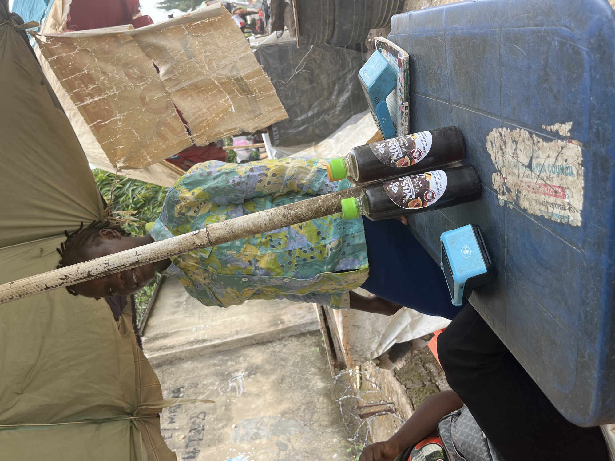 The image depicts a street vendor's stall in Abuja, Nigeria, likely an informal market setting. A woman, presumed to be the vendor, stands partially obscured by a bamboo pole supporting a tattered canvas canopy. She wears a light-colored, patterned shirt and dark blue trousers. In the foreground, on a blue plastic table, are two glass bottles of what appears to be dark liquid, possibly honey or a similar product, labeled "NOS". A blue card reader is placed on a stack of books next to the bottles. A portion of another person's arm and torso, wearing a patterned garment and dark pants, is visible on the left side of the frame, suggesting another individual is present or seated nearby. The background reveals a rough concrete wall with graffiti, some barbed wire, and other stalls or structures characteristic of an outdoor market. The overall scene conveys a busy, informal commercial environment.