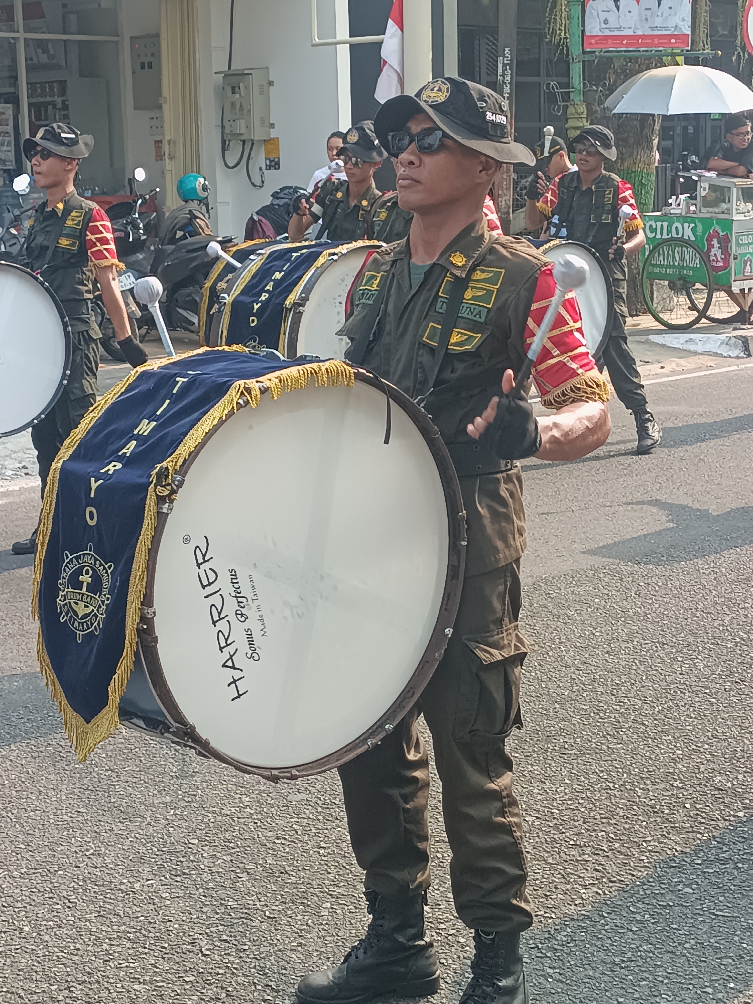 The image depicts a street scene in Sleman, Indonesia, featuring a marching band. In the foreground, a young man, dressed in a dark green military-style uniform with a black baseball cap, sunglasses, and black gloves, plays a large bass drum. The drum is white with the brand name "HARRIER" and "Sonus Perfectus Made in Taiwan" printed on it. A blue banner with gold trim and fringe hangs from the drum, bearing a crest with an anchor and the words "PANA JAYA SAMUD" and "DRUM BAND". Behind him, other band members in similar uniforms can be seen carrying drums. To the left, motorcycles are parked along the side of the street. In the background, buildings and a food cart selling "CILOK" are visible, along with spectators and trees. The lighting suggests it is daytime, with shadows cast on the asphalt road.