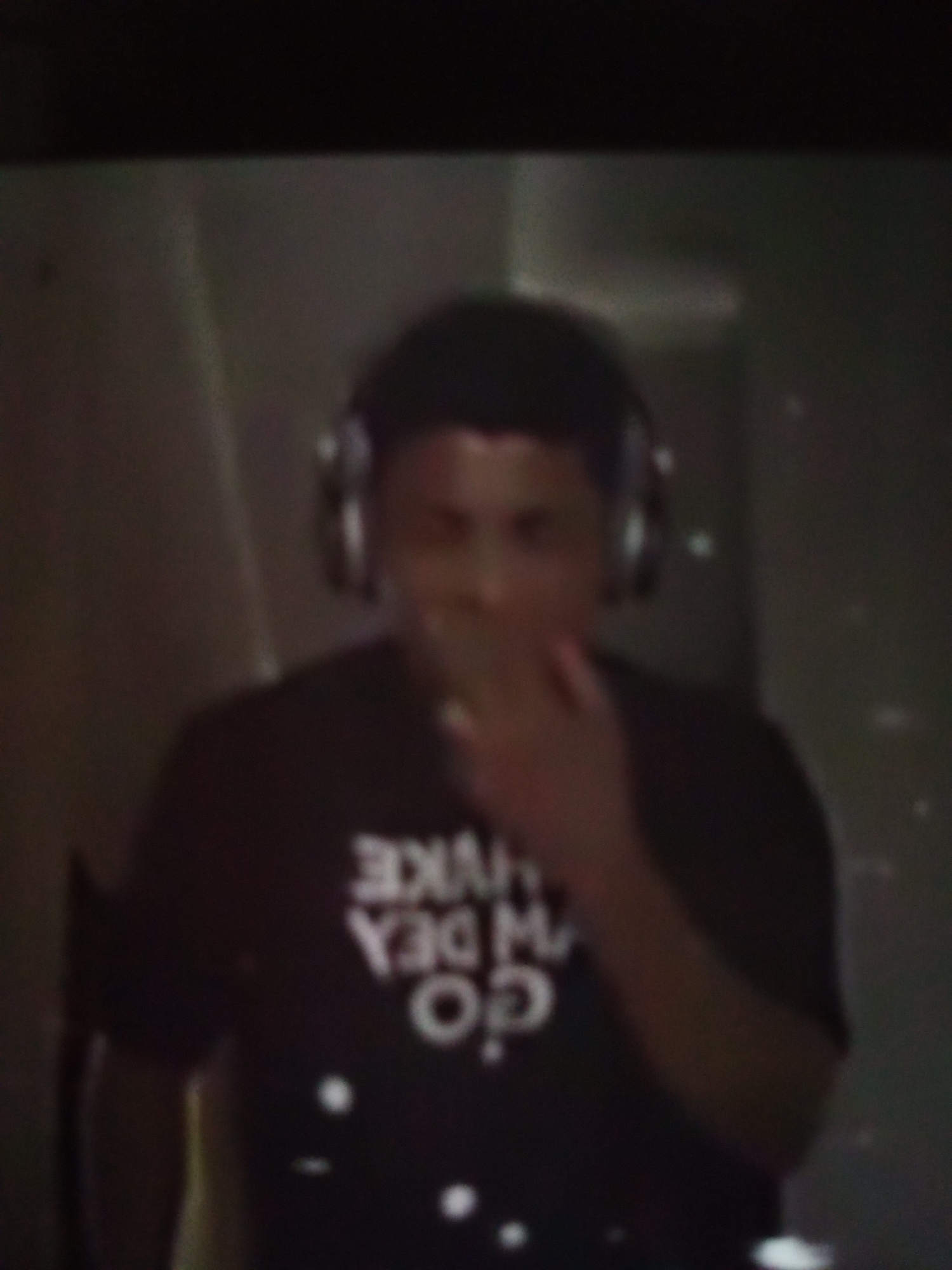 A person wearing headphones and a dark t-shirt with white lettering is partially visible in the frame. The lettering on the t-shirt appears to be in a stylized font and reads "GO DEA MAKE". The individual has their right hand near their mouth. The background is dark and appears to be an indoor environment with some indistinct shapes. There are several blurry white spots scattered throughout the image, potentially indicating a low-light condition or visual artifact. The specific location within Jalingo, Nigeria, cannot be determined from the visual information provided.