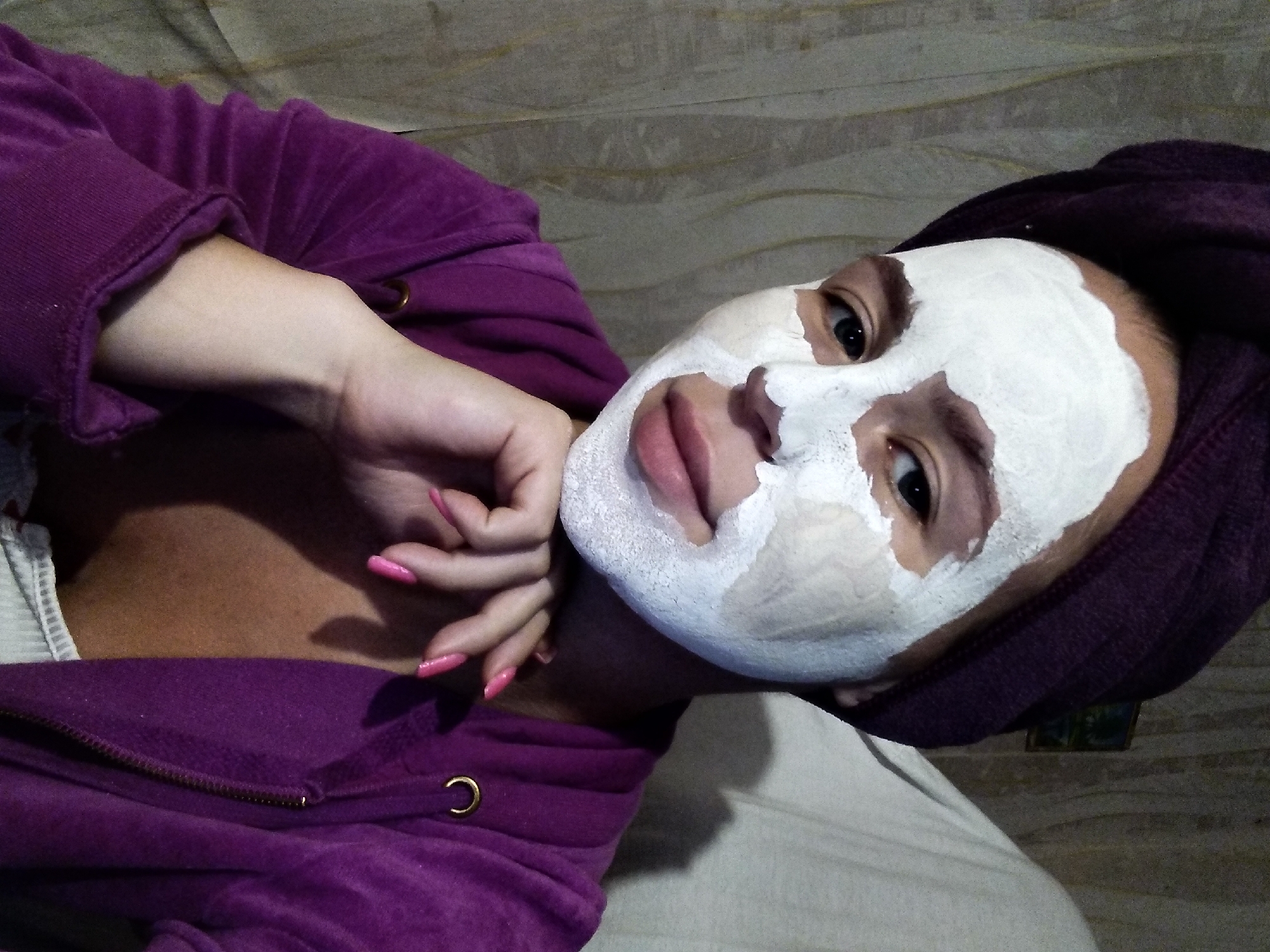 A young woman is pictured from the chest up, wearing a purple towel wrapped around her head and a purple zip-up hoodie. She has a white facial mask applied to her face, covering her nose, cheeks, and forehead, with only her eyes and lips visible. Her right hand is resting on her chin, and her fingernails are painted pink. The background consists of a light-colored, textured curtain and a wall with some faint markings. The lighting is dim, creating shadows on the right side of her face and neck. The image appears to be a selfie taken indoors.