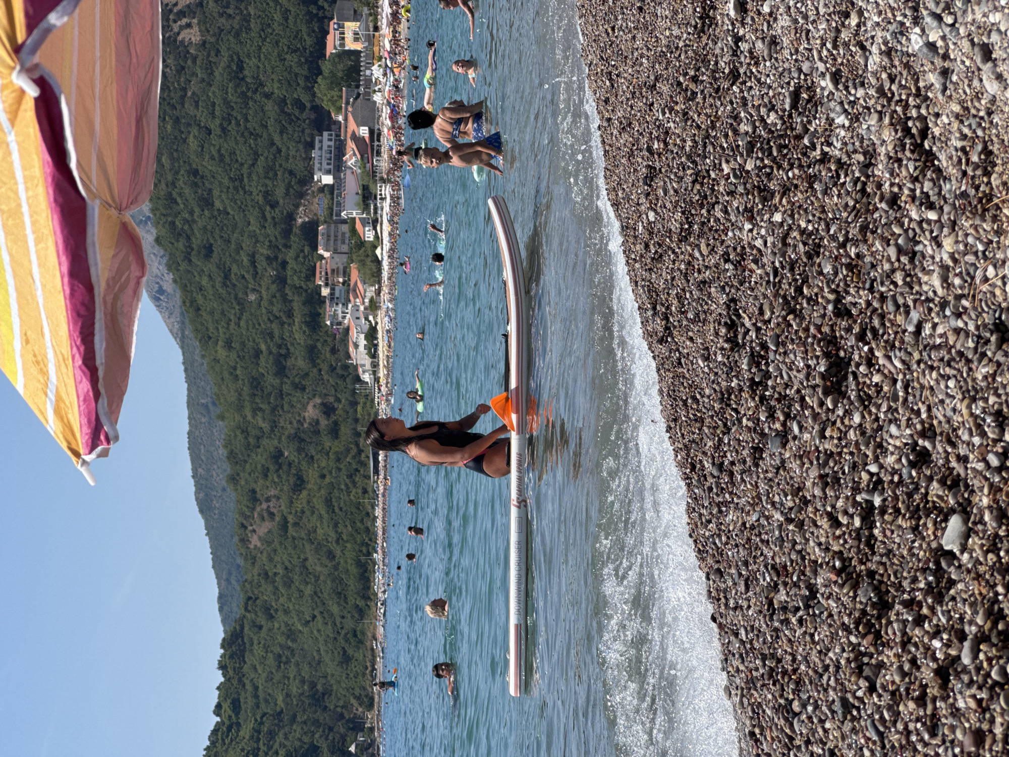 This image captures a sunny day at the beach in Đurovići, Montenegro, with the Adriatic Sea in the foreground and a lush green mountainside dotted with buildings in the background. A person in a black swimsuit is on a stand-up paddleboard, holding an orange paddle. Several other individuals are visible in the water, some swimming and others relaxing. The beach itself is composed of small, smooth pebbles. A large, colorful beach umbrella is partially visible at the top of the frame, casting a shadow over the scene. The water is a clear blue, with small waves lapping at the shore. The general atmosphere is one of leisure and summer recreation.