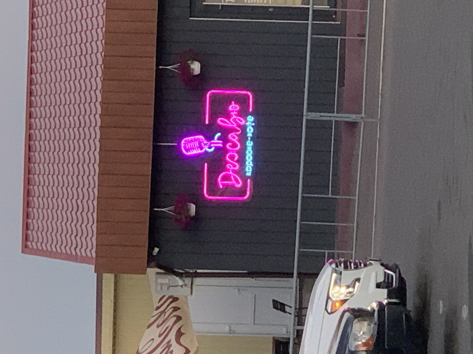 The image depicts the exterior of a building with a neon sign that reads "DescaBio" and below it "караоке-кофе." The neon sign, illuminated in pink, is mounted on a dark-colored facade. Above the sign, two hanging flower baskets are visible, with one containing a plant and the other appearing to have red flowers. The building has a reddish, tiled roof. To the left of the building, a portion of a light-colored awning with dark red lettering is visible. In the foreground, the front section of a white SUV and a dark-colored vehicle are parked. A metal fence runs across the scene in front of the building. The sky above is overcast. The image appears to be taken in Kapyĺ, Belarus, as indicated by the context.