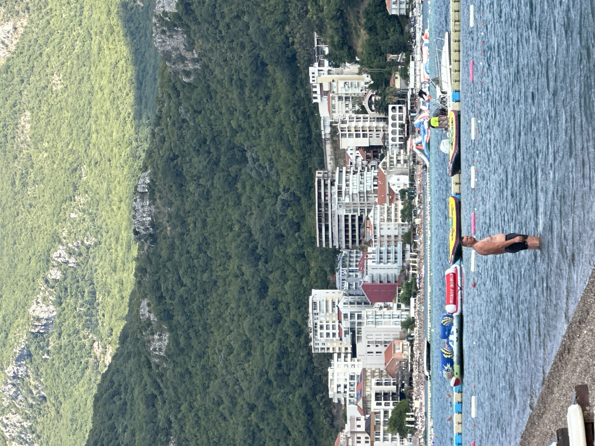 The image captures a scene at the Bura beach in Boreti, Montenegro. In the foreground, a man stands in shallow, blue water, facing towards the left. His body is bare from the waist up, and he is wearing dark swim trunks. The water has gentle ripples, and a concrete shoreline is visible in the lower left corner.
In the mid-ground, several inflatable water toys are anchored in the water. These include a yellow inflatable, a large blue and yellow inflatable with "BG-314 N" visible on its side, and a red inflatable that reads "GREAT EAGLE". A white motorboat is docked near these inflatables. Two individuals, one wearing a yellow shirt and black shorts and another wearing a green shirt and yellow shorts, are interacting with the boat and inflatables. A yellow inflatable tube is also visible to the left of the frame. A line of buoys marks a boundary in the water.
The background features a stretch of beach lined with umbrellas and sunbathers, followed by a dense cluster of white and pastel-colored buildings. These buildings range from low-rise structures to multi-story hotels. Behind the buildings, a steep, densely forested green mountain rises, its upper slopes sparsely dotted with rocky outcrops. The sky is partly cloudy, casting shadows on the mountain.