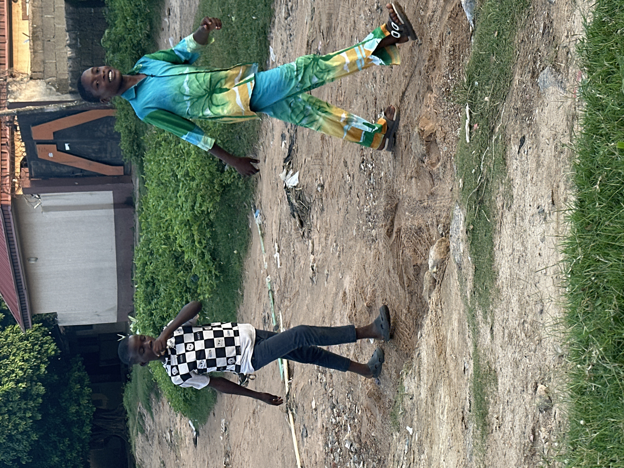 In Narayi, Nigeria, two young boys walk on a dirt road. The boy on the left wears a black and white checkered t-shirt with graphics and dark jeans. He is holding something up to his face. The boy on the right wears a colorful shirt and pants with a tropical print and a pair of sandals. He is smiling and looking towards the viewer. The background shows a building with a red roof, a metal gate, and lush green vegetation. The ground is dusty and uneven, with some scattered debris.