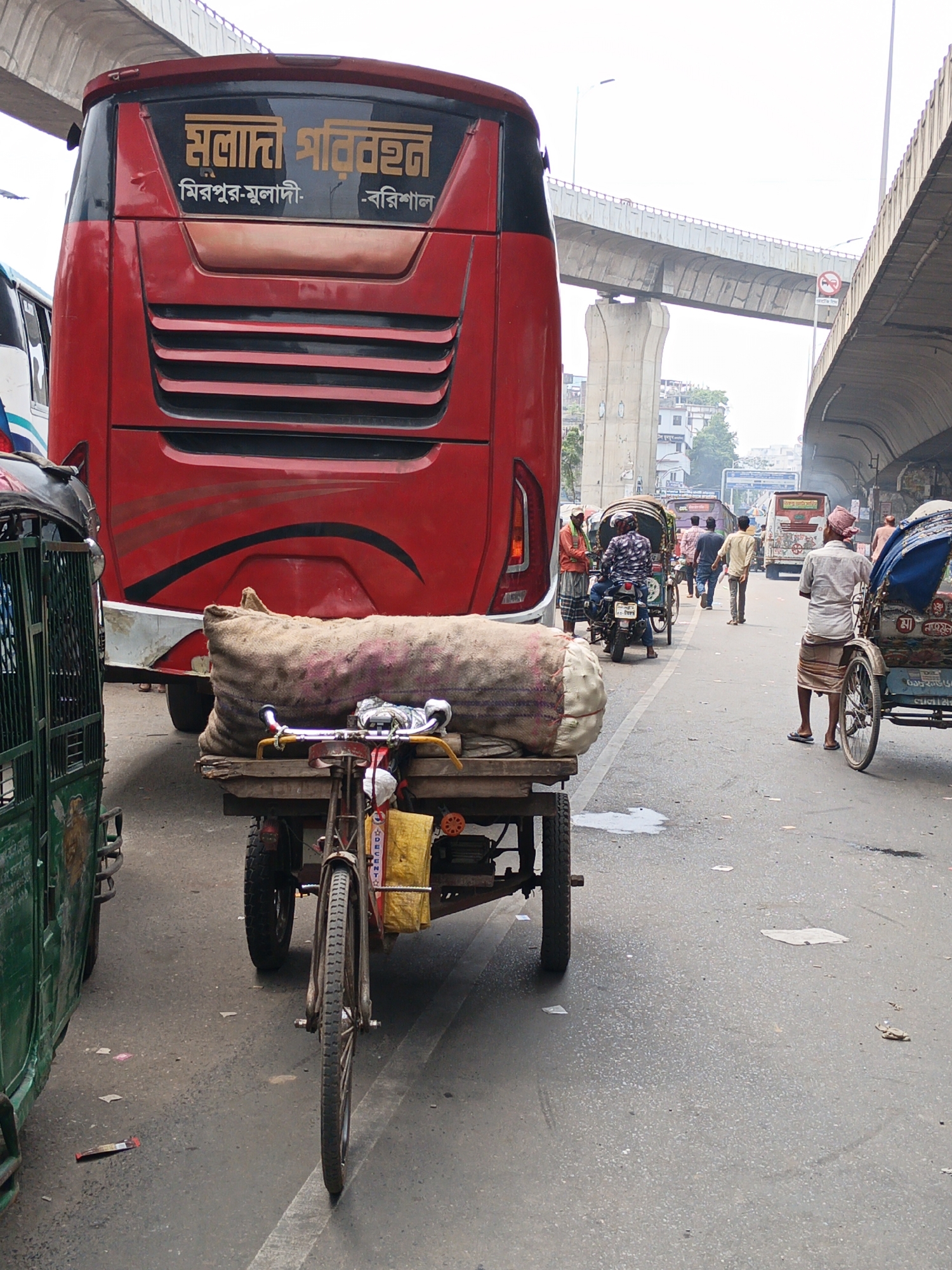 The image shows a busy street scene in Dhaka, Bangladesh, with a red bus dominating the left side of the frame, its rear facing the viewer. The bus has the Bengali words "সোনালী পরিবহন" (Sonali Paribahan) and route destinations written on its back: "মিরপুর-মুলাদি" (Mirpur-Muladi) and "-বরিশাল" (-Barisal).
In front of the bus is a small, bicycle-pulled cart loaded with a large burlap sack, suggesting it is being used for transportation of goods. To the left of the cart is a green auto-rickshaw.
The street is active with people; several men are walking or riding motorbikes further down the road. A rickshaw puller stands on the right side of the frame, ready to transport a passenger. Overhead, a concrete overpass structure spans the street, casting shadows and indicating an urban transportation network. Litter and debris are scattered on the road surface. The overall atmosphere indicates a bustling urban environment typical of Dhaka.