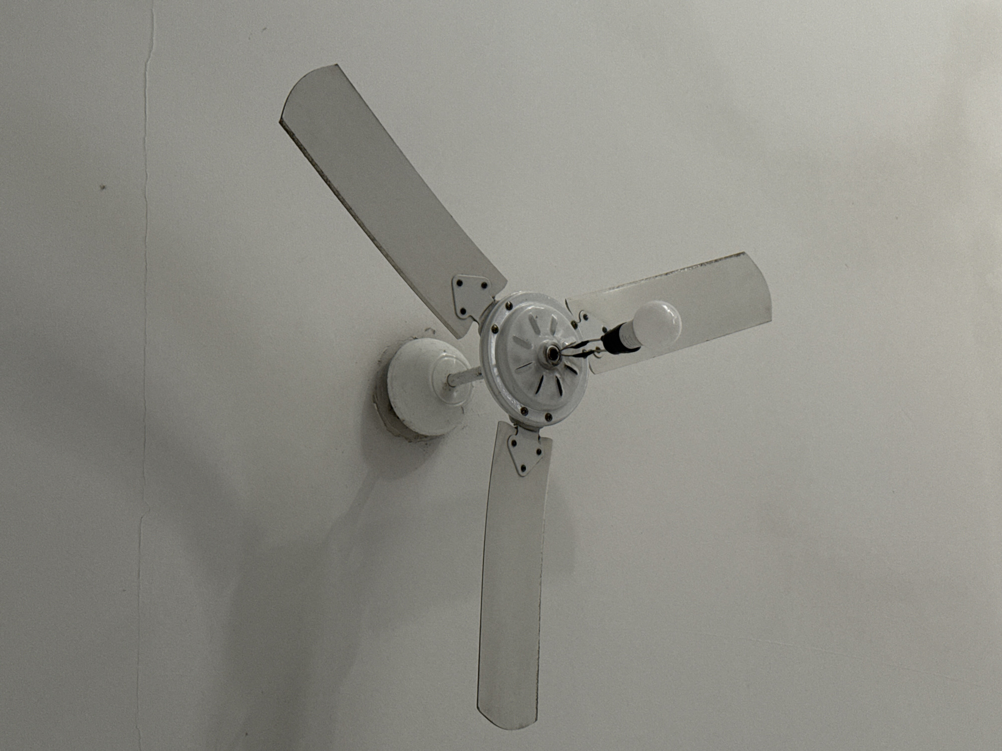 A white ceiling fan with three blades is installed on a white ceiling. A single light bulb hangs from the center of the fan. The ceiling appears to have some minor cracks. The image does not provide any information about the city of Cabo Frio, Brazil.