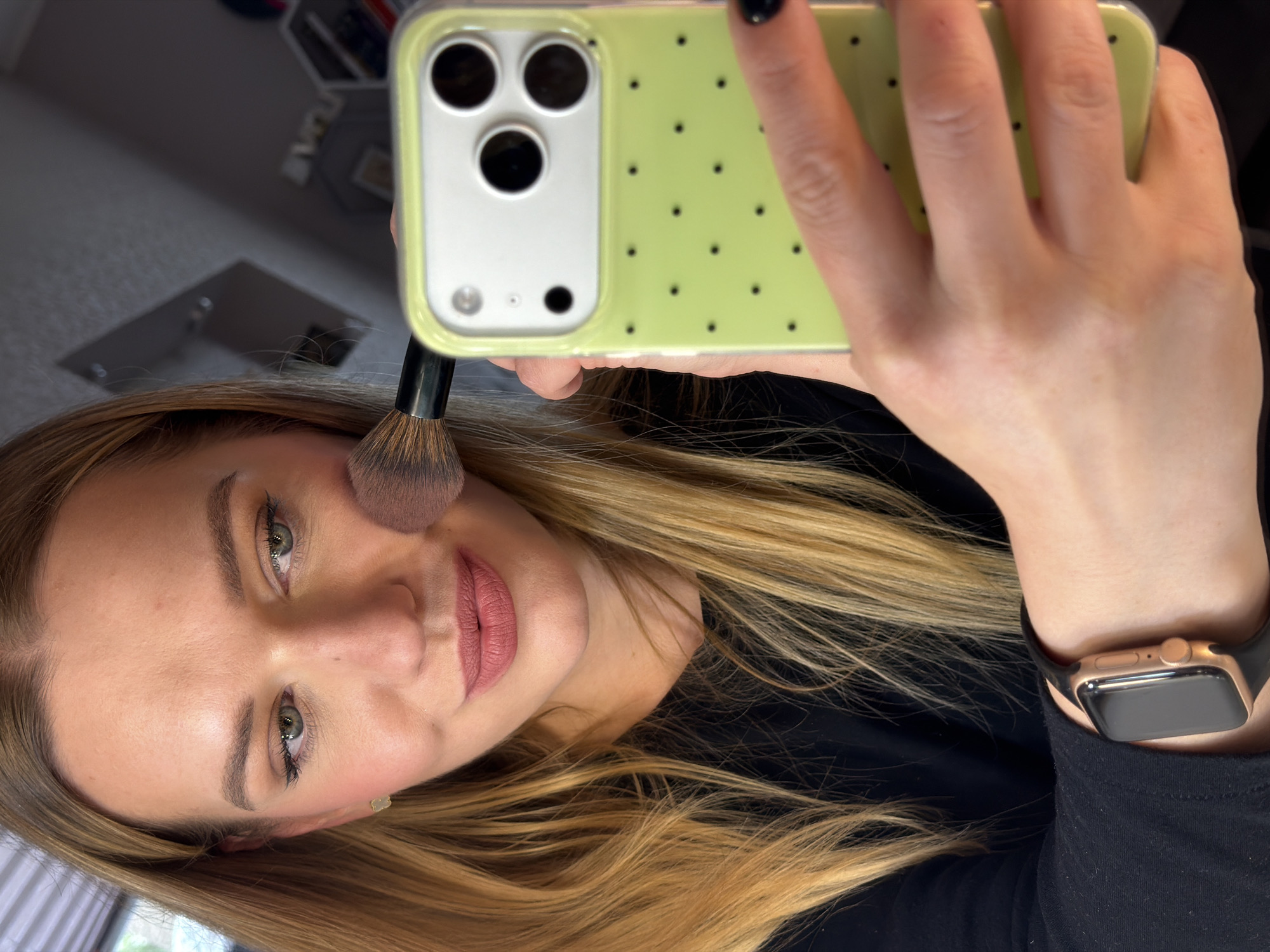 The image shows a woman holding a phone in front of her face. She is applying makeup with a brush. The phone has a light green case with small black dots. The woman is wearing a black shirt and a gold-colored smartwatch with a black band. The background is blurry and does not provide any specific details about the location, making it impossible to determine if it is in Dreiliņi, Latvia.