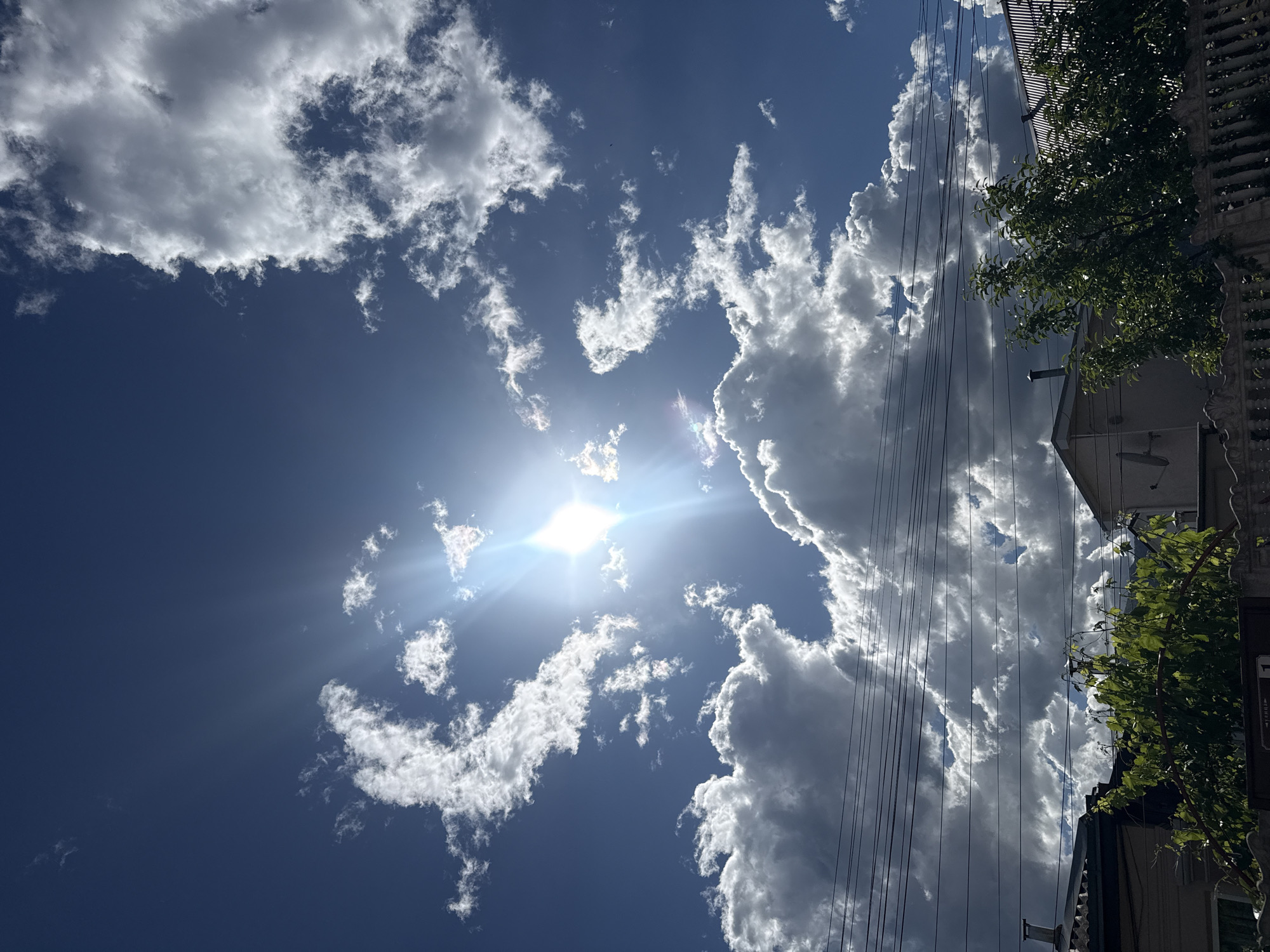 The image shows a daytime sky with scattered cumulus clouds and the sun shining brightly. The sky is blue, with variations in tone due to the cloud cover. The sun's glare is prominent, and its rays extend outwards. On the right side of the image, a portion of a building is visible, with green foliage appearing near its edge. Power lines run vertically through the right side of the frame, intersecting the view of the building and clouds. The location is Unknown, Unknown.