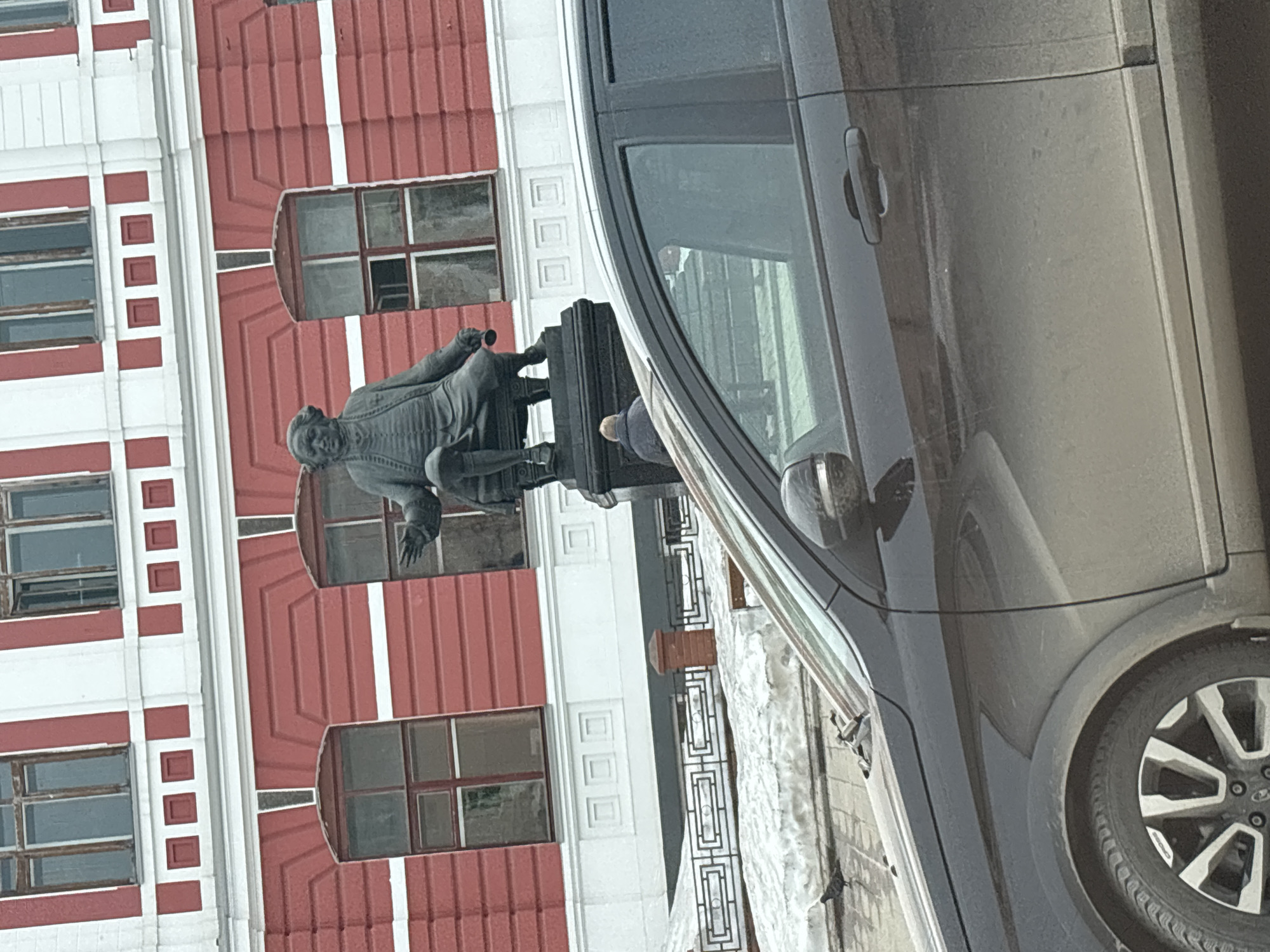 The image shows a statue of Vasily Tatishchev in Perm, Russia. A dark-colored car occupies the right side of the frame, partially obscuring the statue. The statue, set against a red and white building, depicts a seated figure, likely Tatishchev, with one arm extended. A railing with a geometric design is visible in front of the statue, with patches of snow nearby. The building behind the statue features multiple windows with a red and white facade.