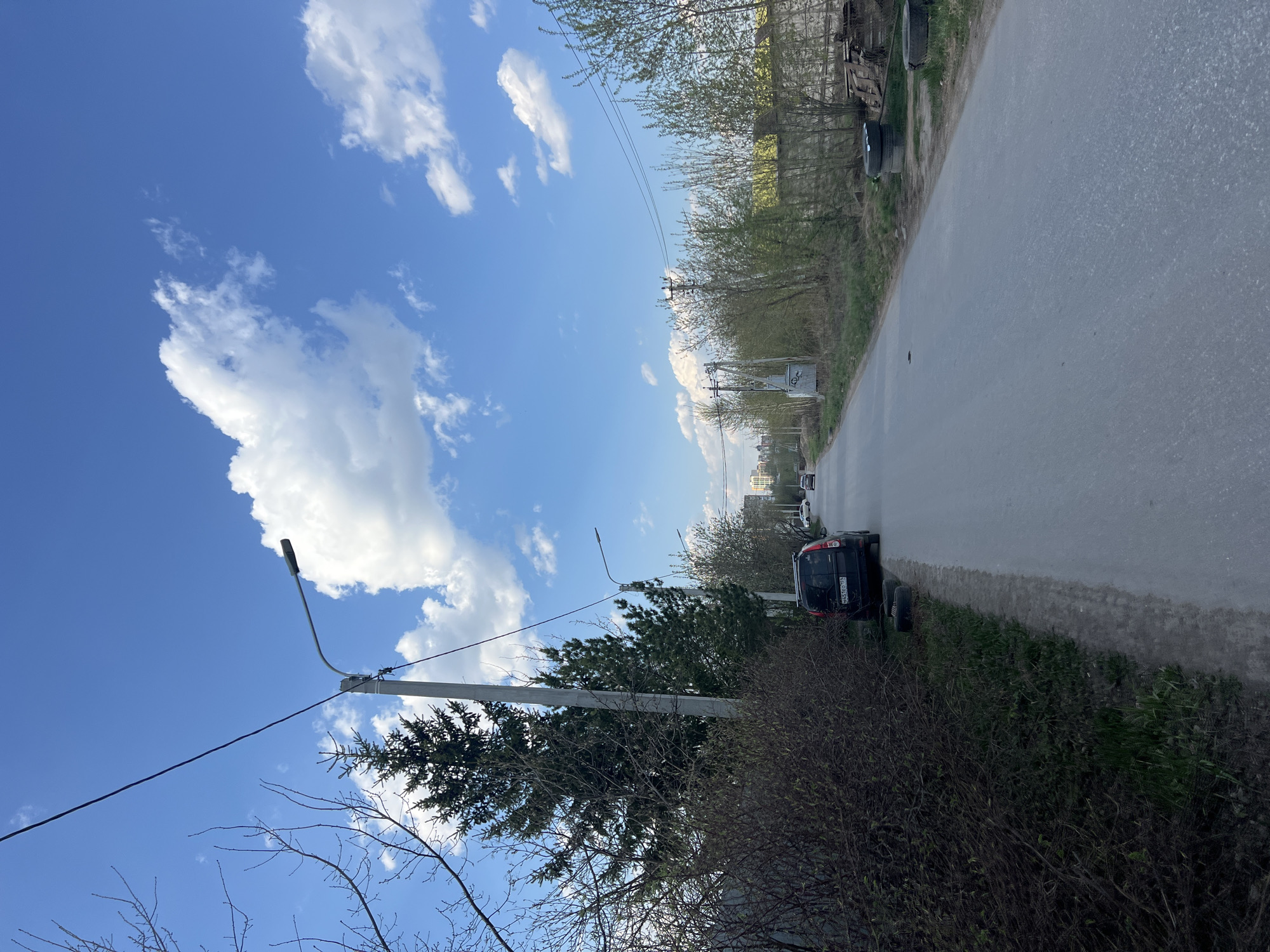 The image depicts a street scene in Perm, Russia, under a bright blue sky with scattered white clouds. The camera is oriented vertically, capturing the edge of a road with a parked dark-colored car and several detached car wheels on the roadside. On the opposite side of the road, there is a line of trees and bushes, partially obscuring a building in the distance. Utility poles with wires are visible, stretching across the sky. A street light extends from a pole on the left side of the road.