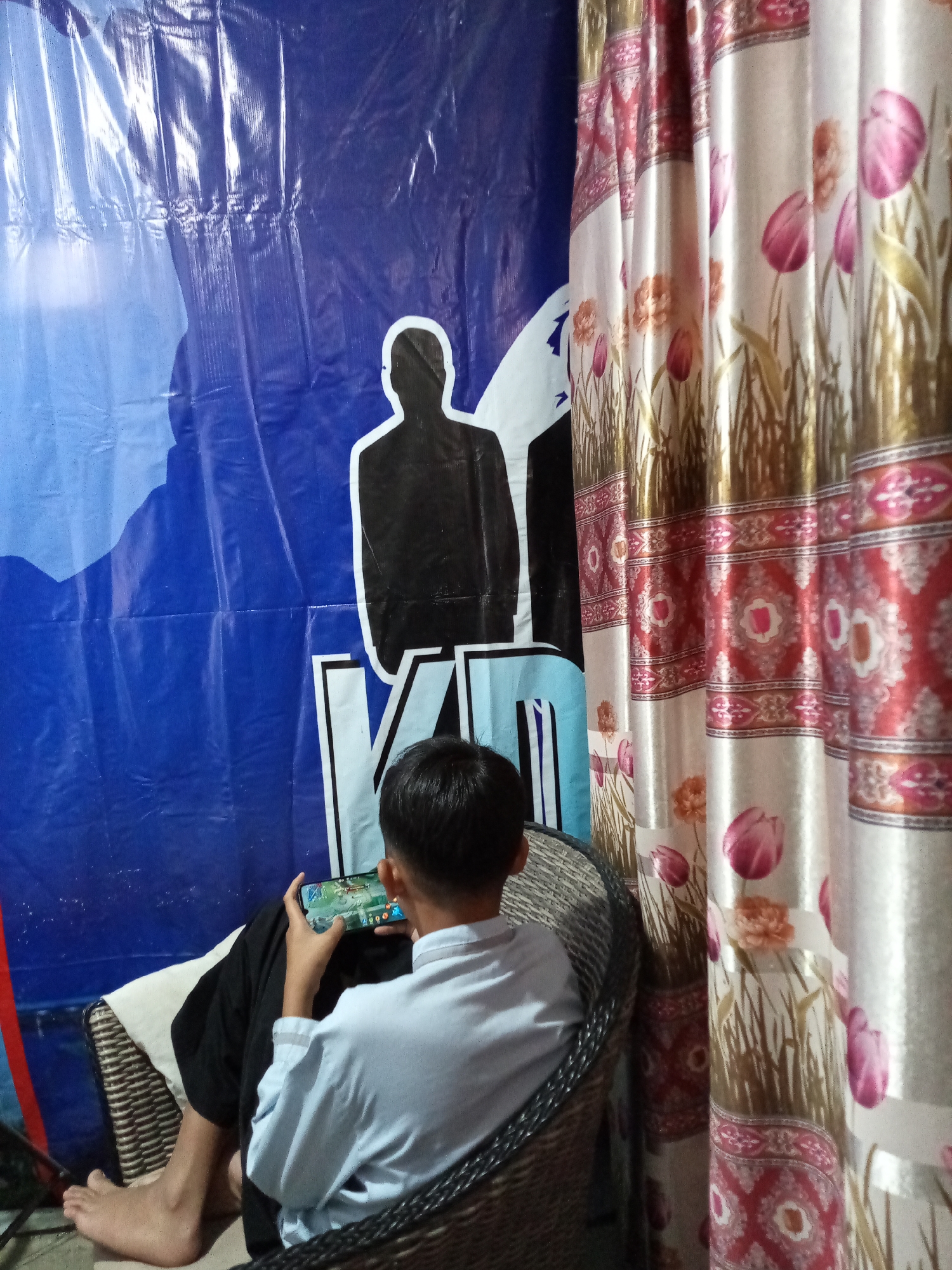 A boy sits in a woven chair playing a mobile game. He is wearing a light blue shirt and dark pants. The background features a blue banner with a silhouette of a person and text, and a patterned curtain with floral designs. The scene appears to be indoors, possibly in a home in Cihanjuang, Indonesia.
