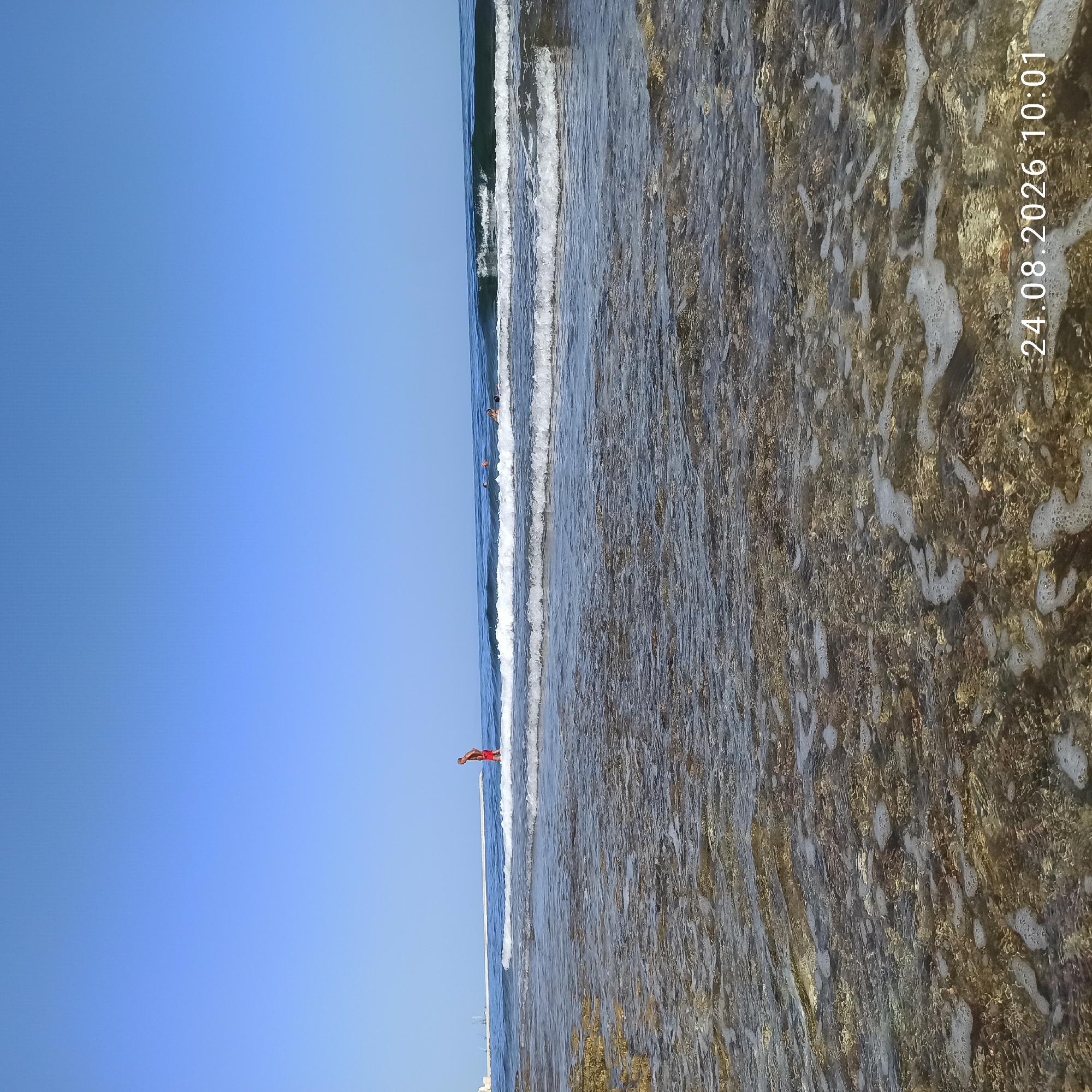 The image depicts a beach scene with shallow, clear water over a rocky seabed. In the distance, several people are visible swimming in the ocean. A single person wearing red shorts stands near the water's edge, facing away from the camera. The sky is clear and blue. A long, man-made structure, possibly a pier or breakwater, extends into the sea on the left side of the image, with some industrial cranes visible near its base. The date "24.08.2026 10:01" is superimposed on the lower right corner of the image. The overall scene suggests a calm, sunny day at a coastal location.