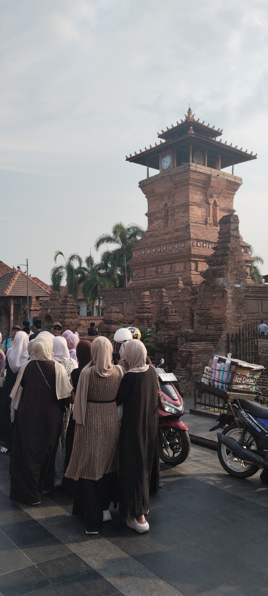 A group of people, mostly women wearing hijabs, stand on a paved area in front of a large brick tower with a clock. The tower has a traditional Javanese architectural style with tiered roofs. To the right of the tower, a cart loaded with stacked cups, likely for ice cream, is attached to a motorcycle. Another motorcycle is parked to the right of the cart. Palm trees are visible in the background. The scene appears to be outdoors, with a light sky. This location is identified as the city of Kauman, Indonesia.