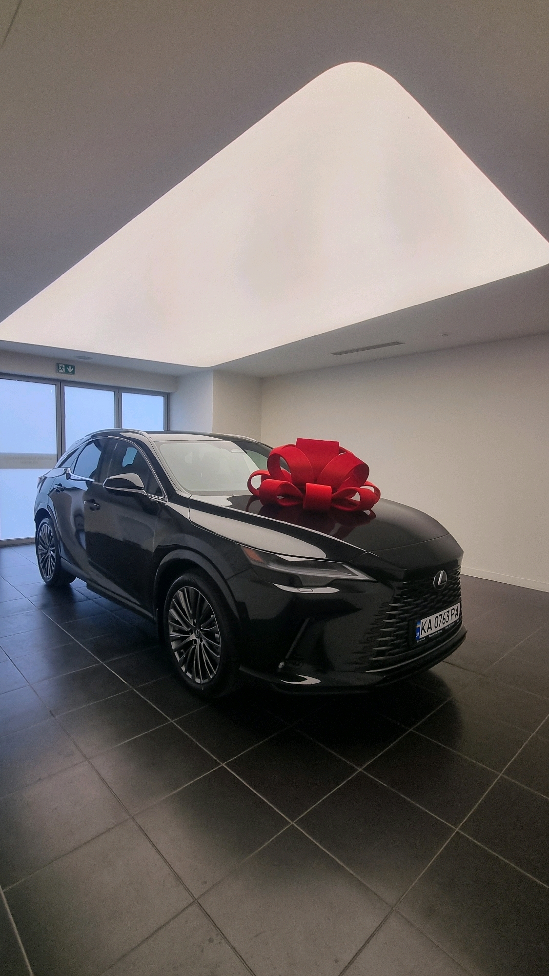 In an indoor showroom, a black Lexus SUV is presented with a large red bow affixed to its hood. The vehicle is positioned on a dark-tiled floor, with large windows visible in the background. Above, a large, illuminated ceiling fixture casts a soft, diffused light. The license plate on the car reads "KA 0763 PA". The overall environment suggests a car dealership setting in Sofiivska Borshchahivka, Ukraine.