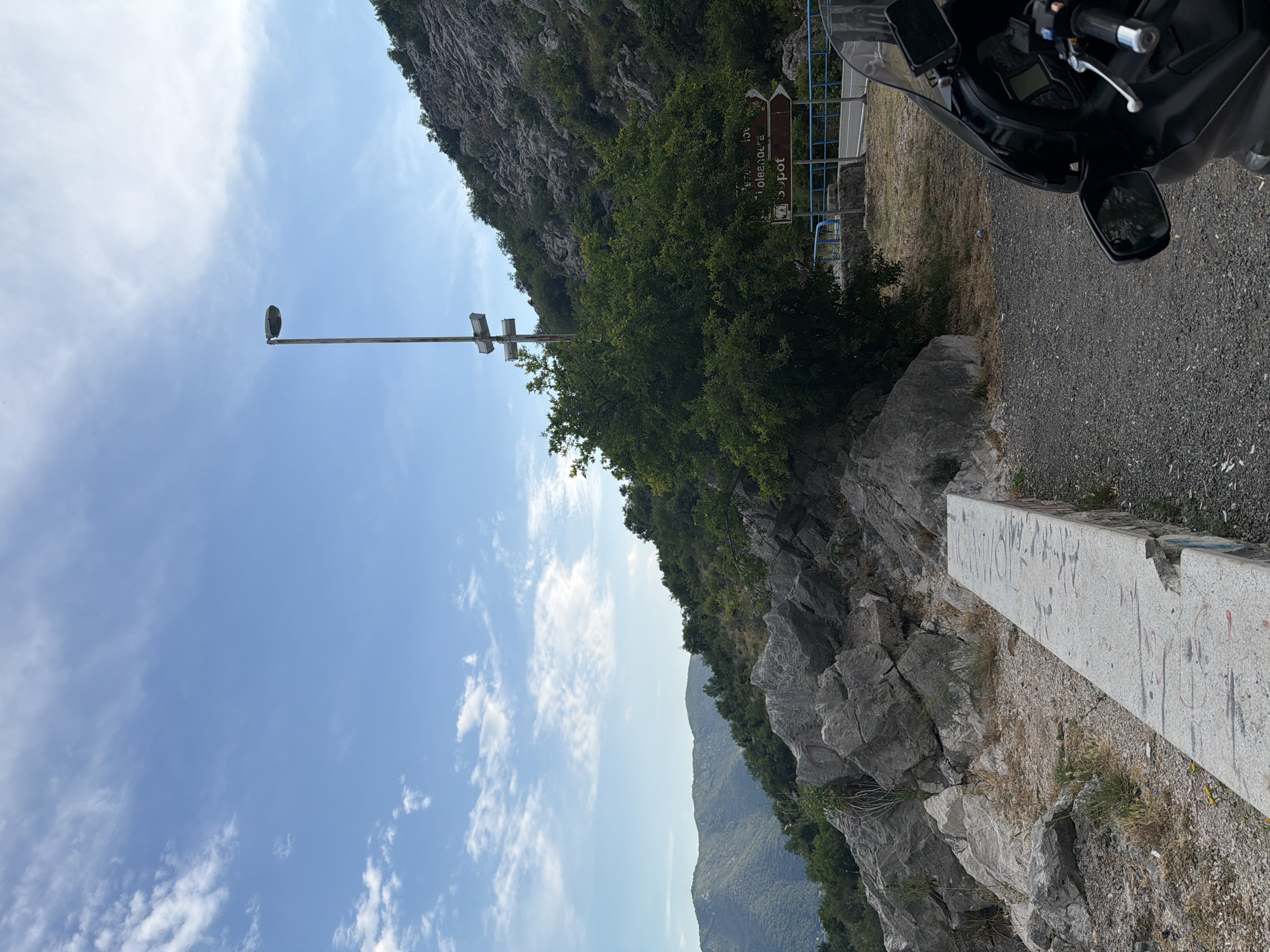 The image captures a scenic view from a roadside lookout point in Risan, Montenegro. In the foreground, the front of a black motorcycle is visible, suggesting a traveler's stop. To the left, a concrete bench with graffiti runs horizontally, leading towards a rocky incline covered in sparse vegetation. The middle ground features a large, leafy green tree and a brown directional signpost indicating "Oplećana" and "Spot". A blue metal railing runs along the edge of the road, separating it from a steep, rocky hillside that dominates the right side of the frame. The background reveals rolling green hills and mountains under a bright blue sky with scattered white clouds. The overall environment is mountainous and coastal, characteristic of the Bay of Kotor.