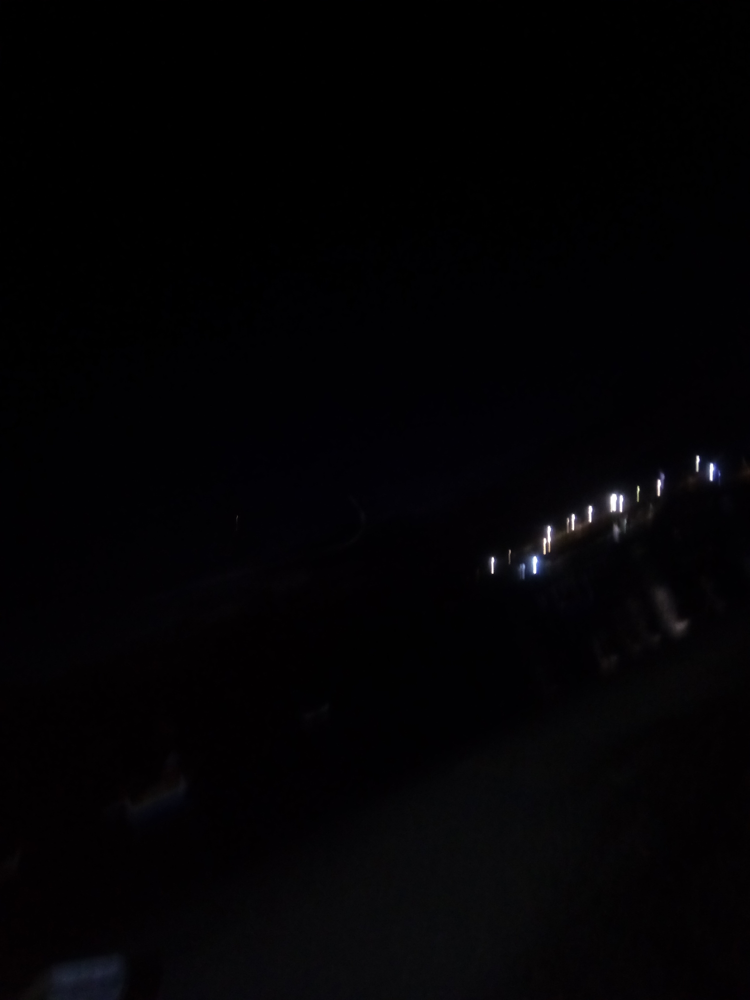 The image appears to be a nighttime scene in Juan Griego, Venezuela. Along a shore line, there are multiple blurred lights, possibly from buildings or streetlights. In the foreground, dark forms suggest land or water. Overall, the image is dark and indistinct.