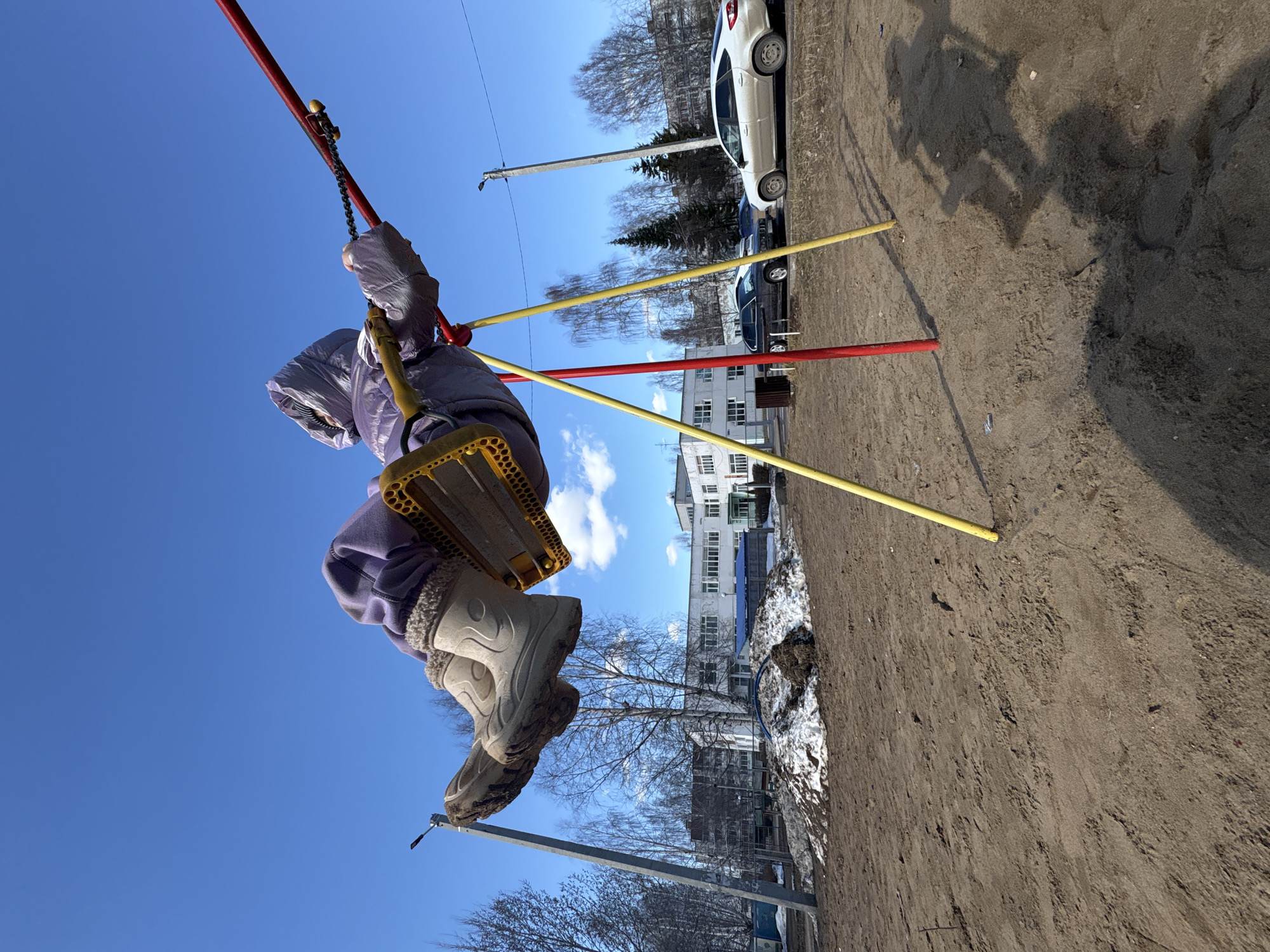 A child in winter clothing is swinging on a playground swing set. The swing set has red and yellow metal supports. In the background, there are apartment buildings, bare trees, and parked cars. There is a patch of snow on the ground near the swing set. The ground around the swing set appears to be dirt or sand. The location is in Chaikovsky, Russia.