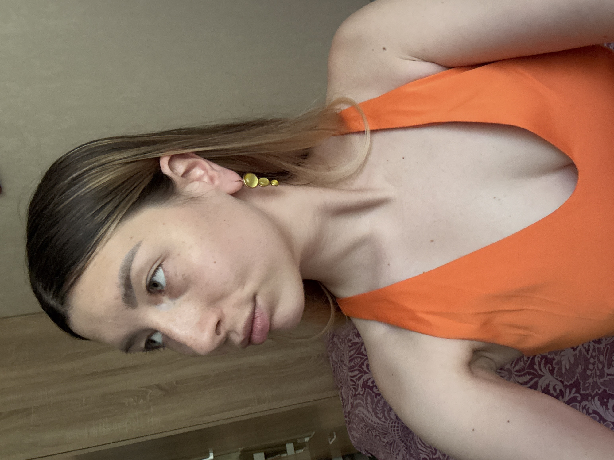 The media file is a close-up, vertically oriented, low-angle photograph of a young woman. She has light skin with freckles, and her dark blonde hair is pulled back. She is wearing an orange garment with a low neckline and a gold earring consisting of three graduated circles. The woman is looking to her left, with her mouth slightly open. Her expression could be interpreted as contemplative or surprised.
The background suggests an indoor setting, possibly a bedroom, with a wooden surface in the lower left and a patterned fabric in the lower right. The lighting appears to be soft and diffused, indicating it could be daytime. No text is visible in the image. The overall mood of the image is intimate and casual. The woman is the sole subject, and there are no other discernible objects or activities taking place. The location context is provided as Moscow, Russia.