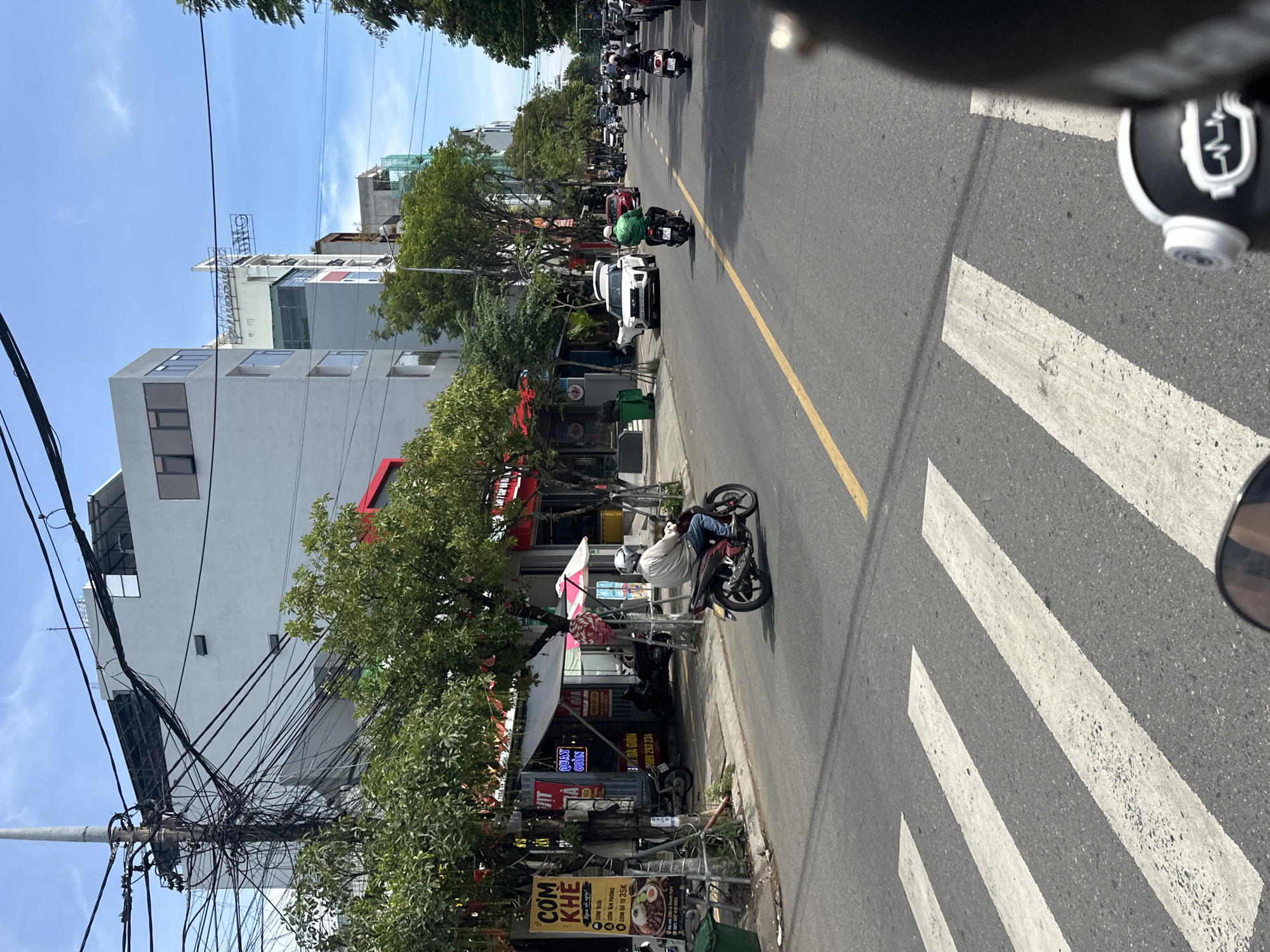 The image depicts a street scene in Da Nang, Vietnam. In the foreground, a person is riding a motorcycle on a paved road marked with a yellow line and a pedestrian crossing. The motorcycle is black and red, and the rider is wearing a light-colored shirt and blue jeans. On the left side of the street, there are several buildings with signs, including one that says "COM KHE". Trees line the sidewalk, and a utility pole with a dense network of wires is visible. In the background, more buildings, other motorcycles, and a white SUV are present. The sky is blue with some clouds. The overall impression is a busy urban environment during daylight.