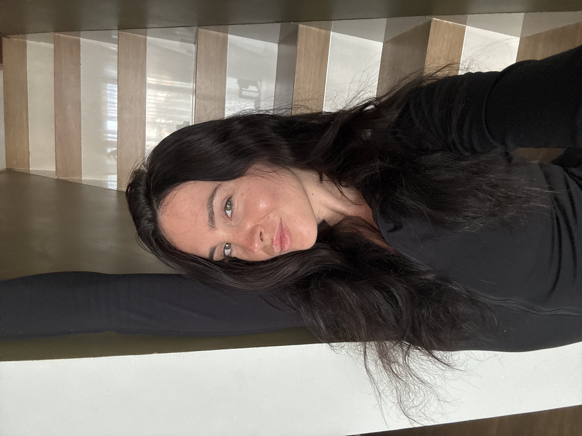 A woman with long, dark, wavy hair is captured from the chest up, facing forward and looking slightly to her left. She is wearing a black, zip-up top. Her left arm is raised and bent behind her head, with her hand touching her hair. Her right arm is not visible. She has light skin with freckles and is looking at the camera with her mouth slightly puckered.
Behind her, a staircase with alternating light wood and white steps ascends. The wall to her left is a plain, light color, and the wall to her right is a dark, muted color. The lighting in the image is soft, casting a warm glow on the woman's face. The overall environment suggests an indoor residential setting. The image does not provide any specific details about the city of North Kuta, Indonesia.