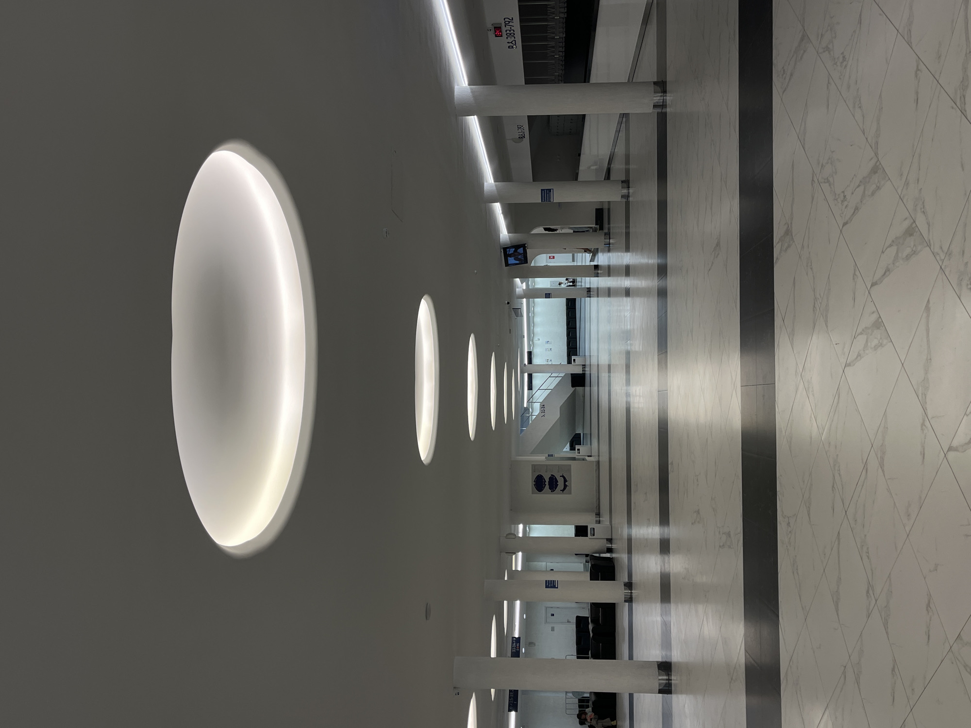 The image shows a wide, modern interior hallway with a white, marble-patterned floor. Several large, white, cylindrical pillars support a white ceiling with recessed circular lighting fixtures. In the background, a staircase is visible, leading upwards. The hallway is clean and well-lit, with a minimalist aesthetic. There are some seating areas with dark couches visible on the left side, and a television screen mounted on a wall further down the hallway. There are a few people present, but they are not the main focus of the image. The overall impression is one of a public building, possibly an airport, train station, or administrative center.