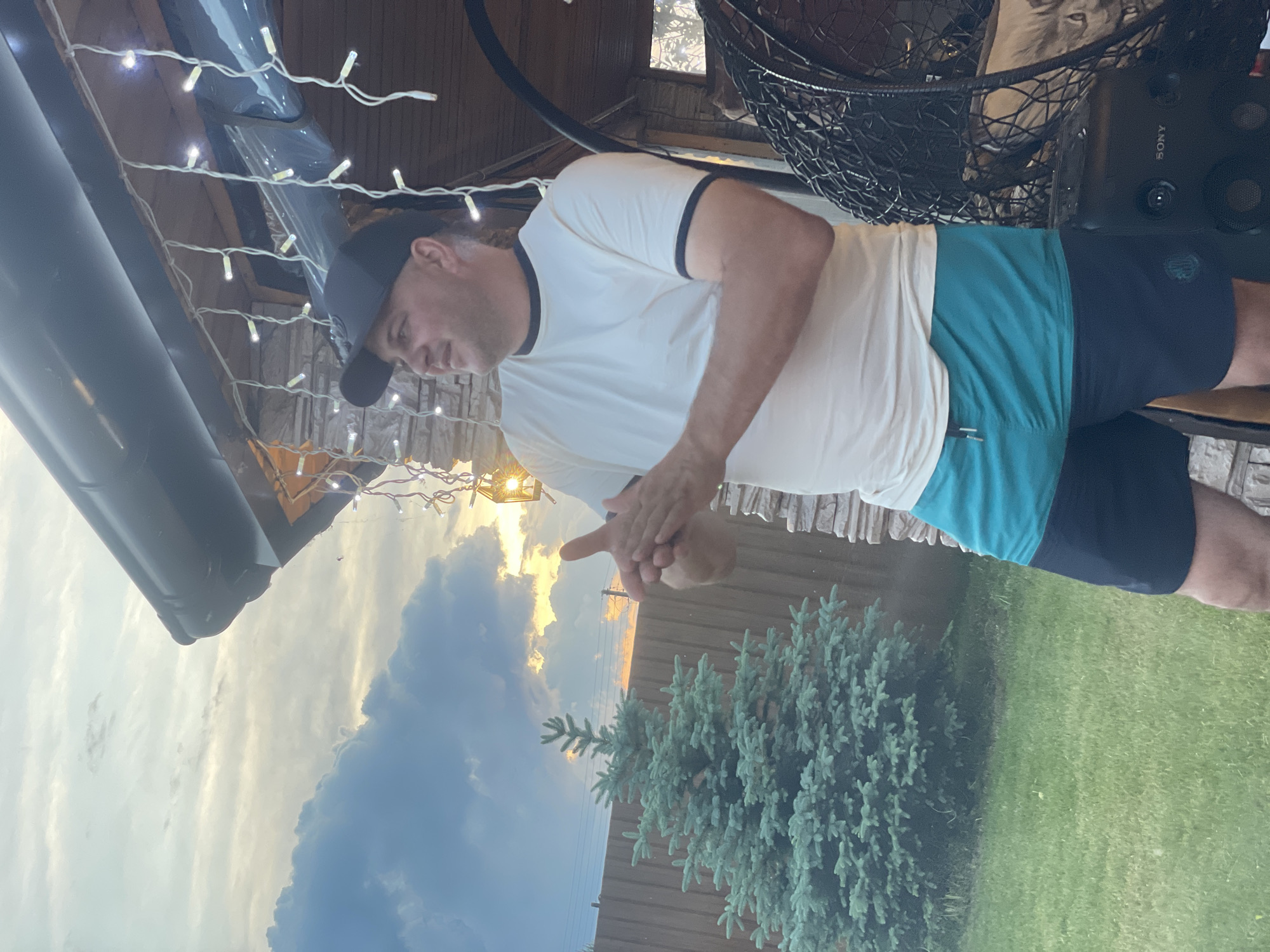 A man in a white t-shirt, black baseball cap, and teal and black shorts stands outdoors in what appears to be a backyard. He is clapping his hands. Behind him, a small evergreen tree stands in a grassy yard, bordered by a wooden fence. A string of lights hangs from the eaves of a structure behind the man, which is made of stone and wood. In the background, the sky is cloudy with hints of sunset colors. The scene is located in Chulym, Russia.
