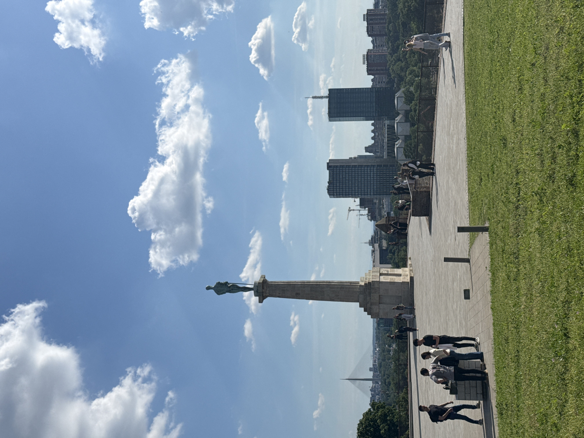 The image depicts the Victor Monument in Belgrade, Serbia, against a backdrop of a blue sky with scattered clouds and the city skyline. The monument, featuring a bronze statue of a nude male figure holding a sword and a falcon, stands prominently on a tall pillar. The Sava River and the Ada Bridge are visible in the background.
On the right side of the image, a paved walkway is visible with a grassy area bordering it. Several people are present on the walkway, appearing to be tourists or visitors. They are standing and walking, some seemingly interacting with the monument or taking pictures. The clothing of the people is casual, suggesting a relaxed, daytime setting. The overall environment suggests a popular tourist destination on a sunny day.