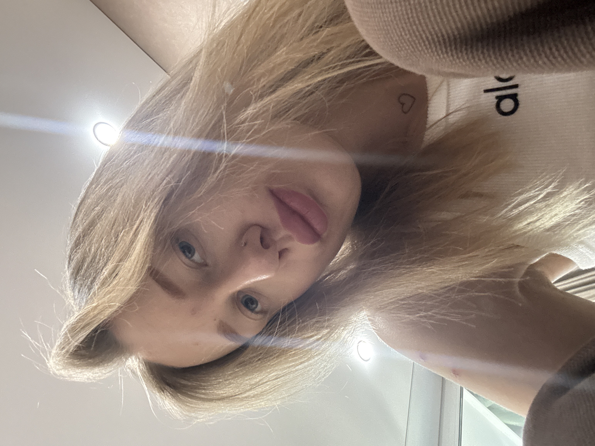 A selfie of a blonde woman is angled downwards. She has blue eyes and her lips are slightly parted. She is wearing a light-colored top with the word "Alo" visible. A small heart tattoo is on her neck. The background is a blurred combination of light and dark surfaces. The location and the city of Ovoshchnoy, Russia are not identifiable from the image.