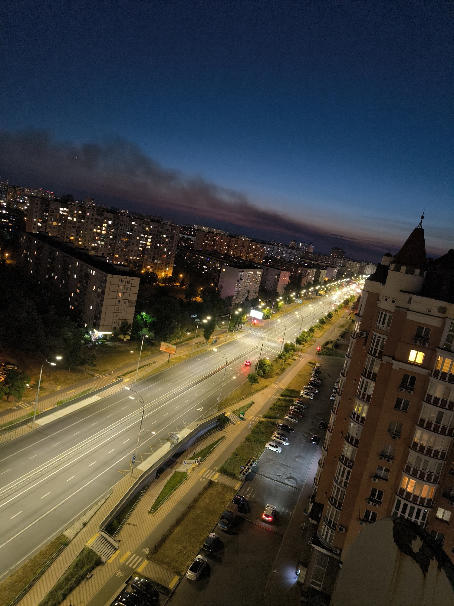 This is a nighttime, high-angle, full shot of a wide, multi-lane road in Kyiv, Ukraine. The sky is a deep twilight blue, transitioning to a darker hue near the horizon, with some wispy clouds tinged pink and orange from the fading light. Smoke or haze is visible in the distance, possibly from a fire.
On the left side of the road, numerous apartment buildings, typical of Soviet-era architecture, are lit up with many illuminated windows, indicating residential occupancy. Trees line the median of the road and the sidewalks, their foliage dark against the artificial lights. Streetlights illuminate the road, casting long shadows and highlighting the white lane markings. Traffic is light, with a few cars visible, some with their headlights on. One red car is seen with its brake lights illuminated.
On the right side of the road, a distinctive building with a pointed, turret-like roof stands out. It's an apartment building with a reddish-orange facade and numerous balconies, many with illuminated windows. The building's architecture suggests a more modern or stylized design compared to the adjacent structures. Parked cars are visible along the side of the road in front of this building.
The overall scene depicts an urban environment at dusk or early night, with the artificial lights of the city creating a distinct atmosphere. The perspective emphasizes the scale of the road and the density of the residential buildings.