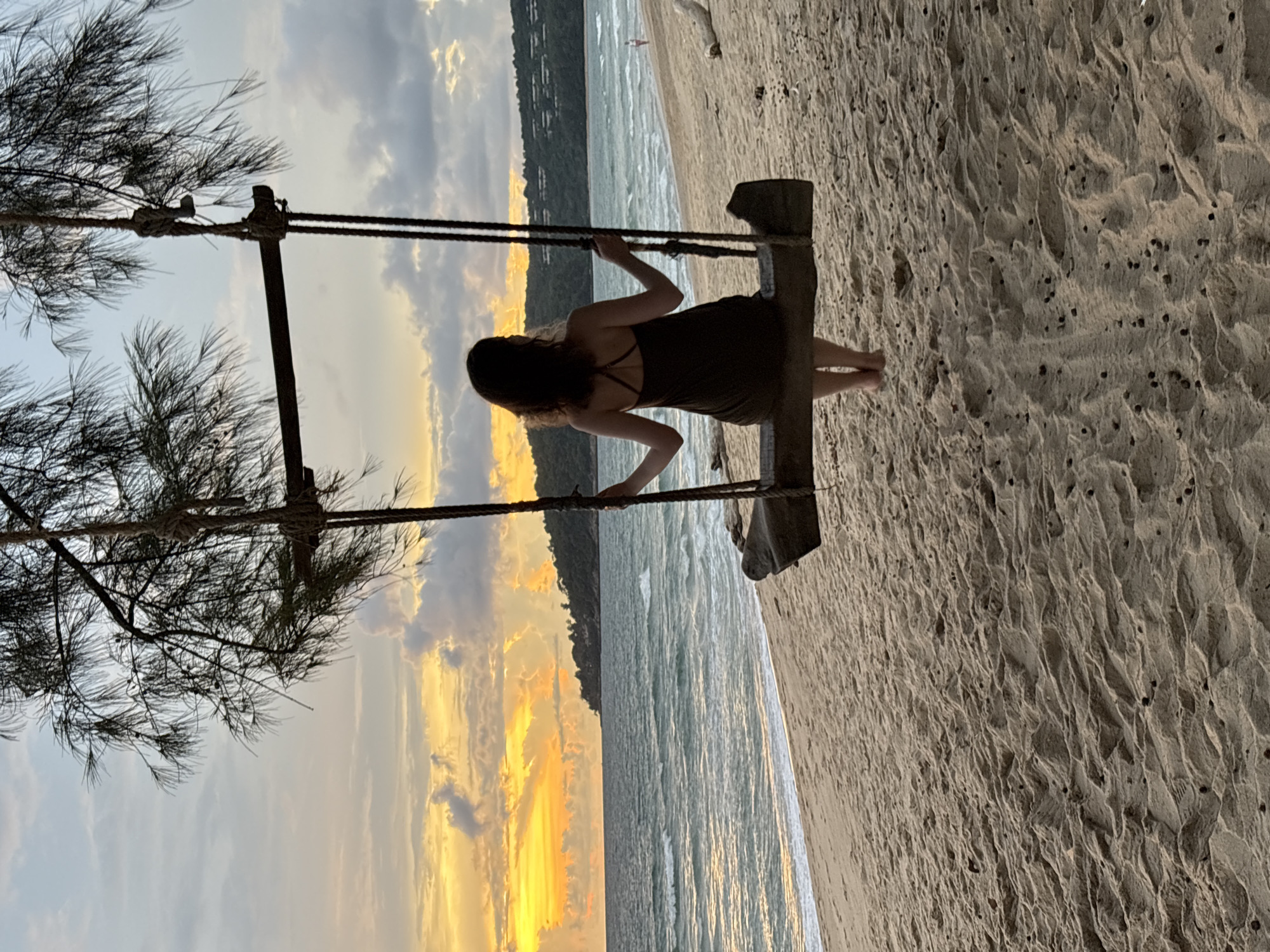 A woman is sitting on a wooden swing on a sandy beach in Choeng Thale, Thailand, facing the ocean at sunset. The sky is filled with dramatic clouds illuminated by the setting sun, casting a warm golden glow over the scene. The ocean waves are gentle, rolling onto the shore. In the background, a tree with thin branches hangs over the swing, its ropes tied to the branches. A distant island with lush vegetation is visible on the horizon. The beach is largely empty, with only a few scattered pieces of driftwood and a single person walking in the distance. The image captures a serene and picturesque moment of a woman enjoying the tranquility of the beach at dusk.
