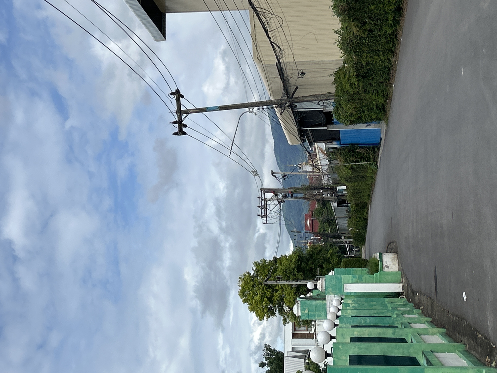 This is a vertical, eye-level, medium-shot photograph taken on a cloudy day in Chang Phueak, Thailand. The scene depicts a paved road curving to the left, bordered on the left by a low, green fence with white spherical finials. Beyond the fence, glimpses of white buildings and lush green foliage are visible. On the right side of the road, a light beige industrial building dominates the foreground, with a dense hedge of green bushes lining its base. A prominent wooden utility pole with numerous black wires stretches across the upper right portion of the frame, converging towards the industrial building. Another utility pole is visible further down the road on the right, also laden with wires. In the distance, a range of green hills is shrouded in haze, with a few scattered red-tiled roofs of other buildings visible amidst the vegetation and power lines. The sky above is a mix of pale blue and heavy grey clouds. There are no people or vehicles visible in the image.