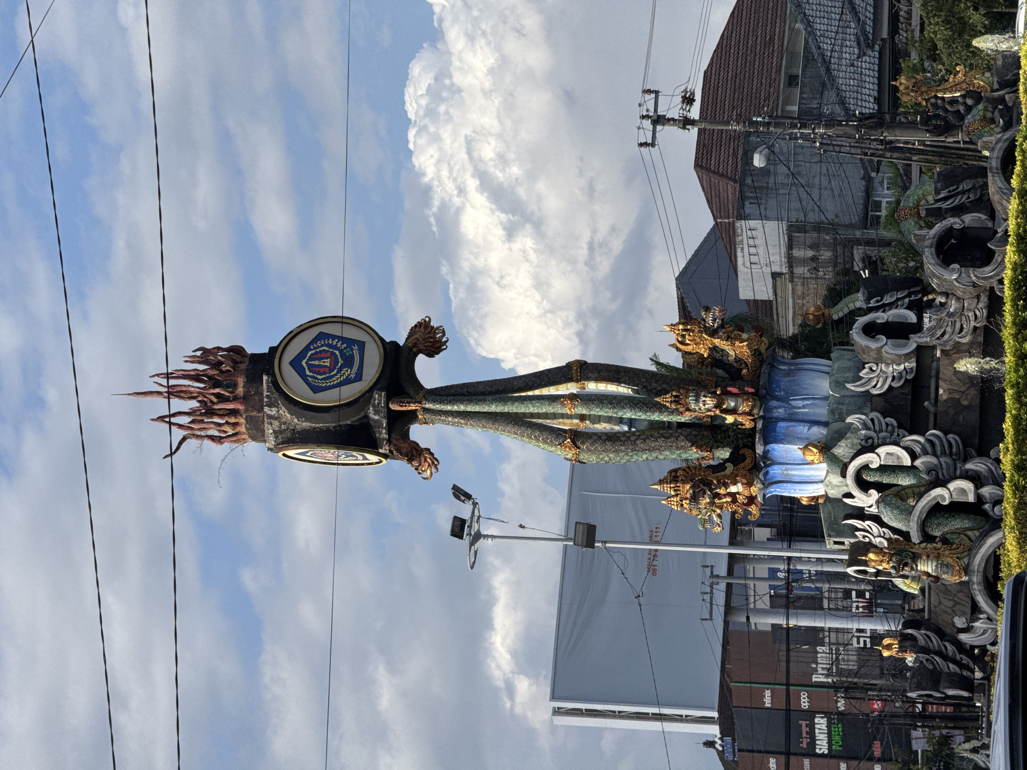 The image shows a monument in Denpasar, Indonesia, featuring intricate carvings of dragons and mythical creatures. The monument is topped with a circular emblem and what appears to be fiery embellishments. The sky above is partially cloudy, with a bright blue expanse visible. In the background, various buildings, utility poles, and a large billboard can be seen, suggesting an urban environment. The monument itself is elaborately decorated, with multiple levels and figures, some of which are gilded. The overall scene depicts a vibrant cultural or historical landmark within a bustling cityscape.