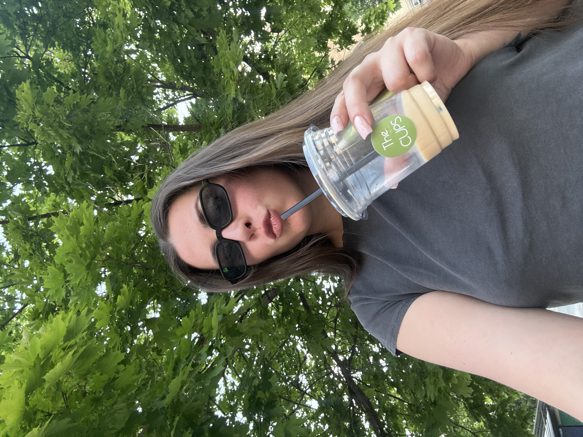 A young Caucasian woman with long brown hair and sunglasses is drinking from a clear plastic cup with a straw. She is wearing a dark gray t-shirt. The cup has a green circular sticker with white text that reads "The CUPS". She is holding the cup with her right hand. The background is filled with lush green trees and dappled sunlight. The scene appears to be outdoors, possibly in a park or on a street with trees.