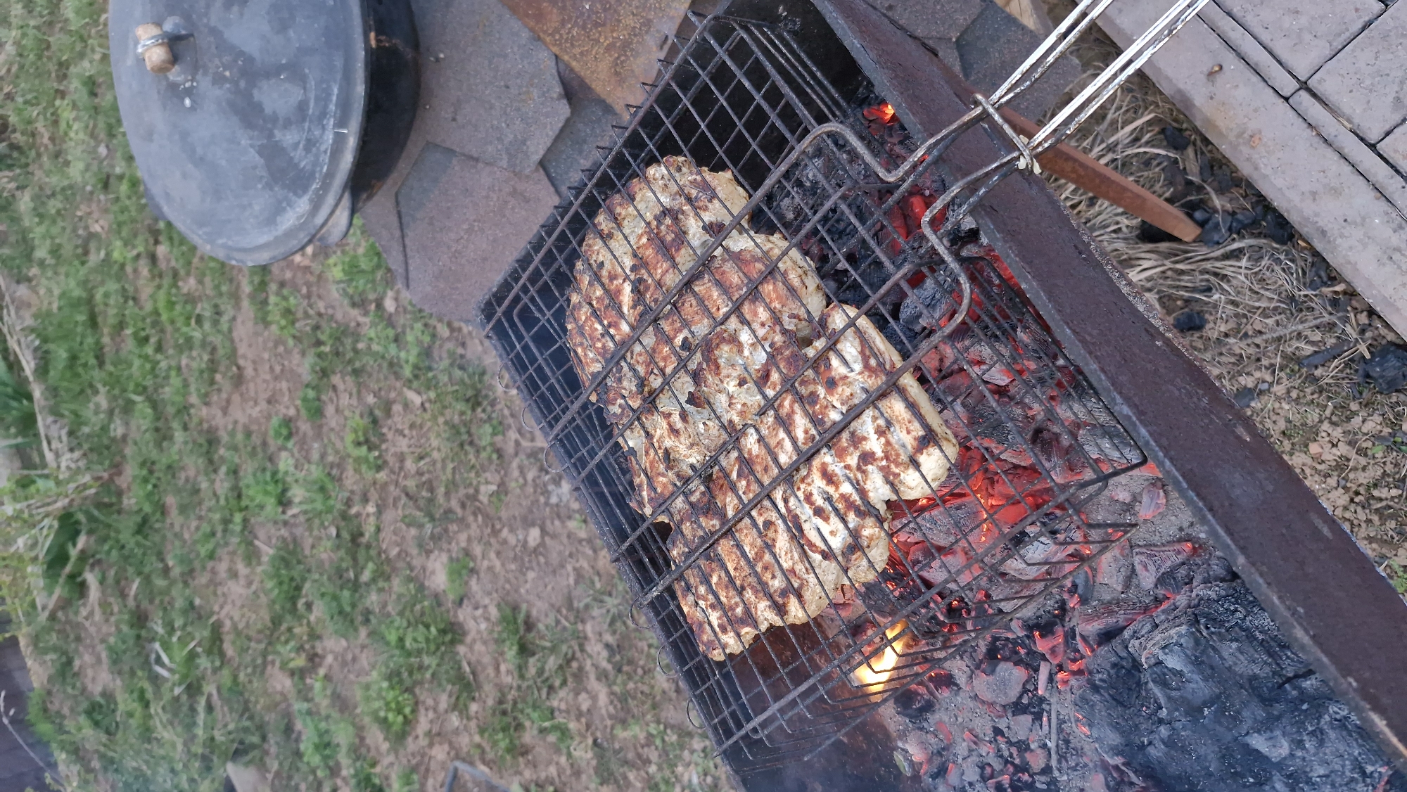 The image depicts food grilling over a charcoal grill, with the coals glowing red. The food appears to be pieces of grilled chicken, marked with grill lines. To the left of the grill is a cast iron pot with a lid, sitting on paving stones, next to a patch of green grass. The location is Otchor, Russia.