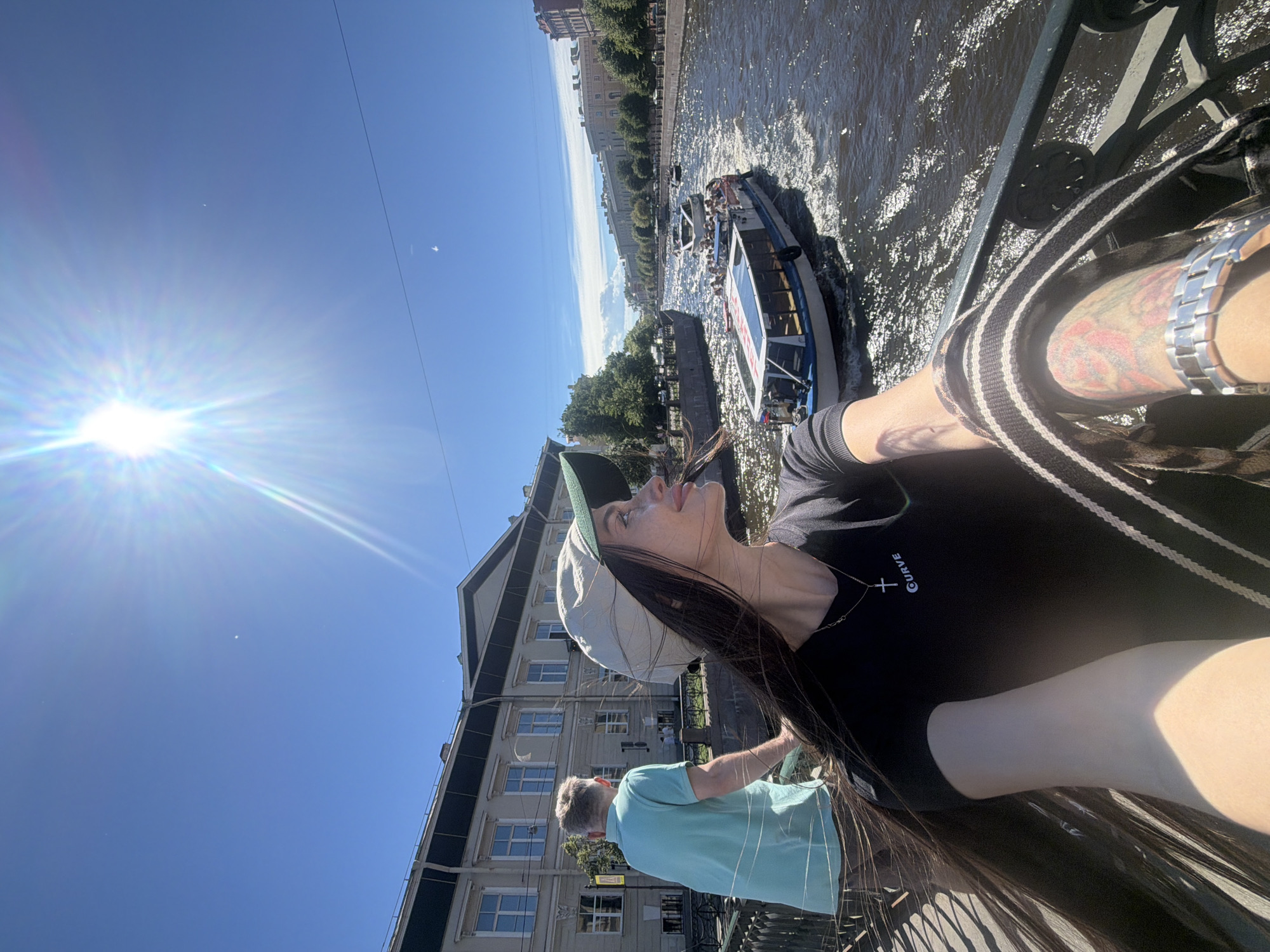 On a sunny day, a woman in a black t-shirt and a light-colored baseball cap takes a selfie on a bridge overlooking a canal in Saint Petersburg, Russia. Her long dark hair is blowing in the wind. She is wearing a silver necklace with a cross pendant and the word "CURVE" below it. Her left arm, adorned with a bracelet and tattoos, is extended, holding the camera. Beside her, a man in a light blue polo shirt stands facing away from the camera, looking towards the canal.
The canal is bustling with activity. Several tour boats, filled with passengers, are navigating the waterway. The water's surface is choppy, reflecting the bright sunlight. On the far bank of the canal, traditional St. Petersburg architecture, characterized by multi-story buildings with large windows, lines the embankment. Green trees are interspersed between the buildings. The sky is clear blue with a few wispy clouds. The sun is high in the sky, casting strong shadows and creating lens flare in the image. The scene captures a vibrant summer day in the city.