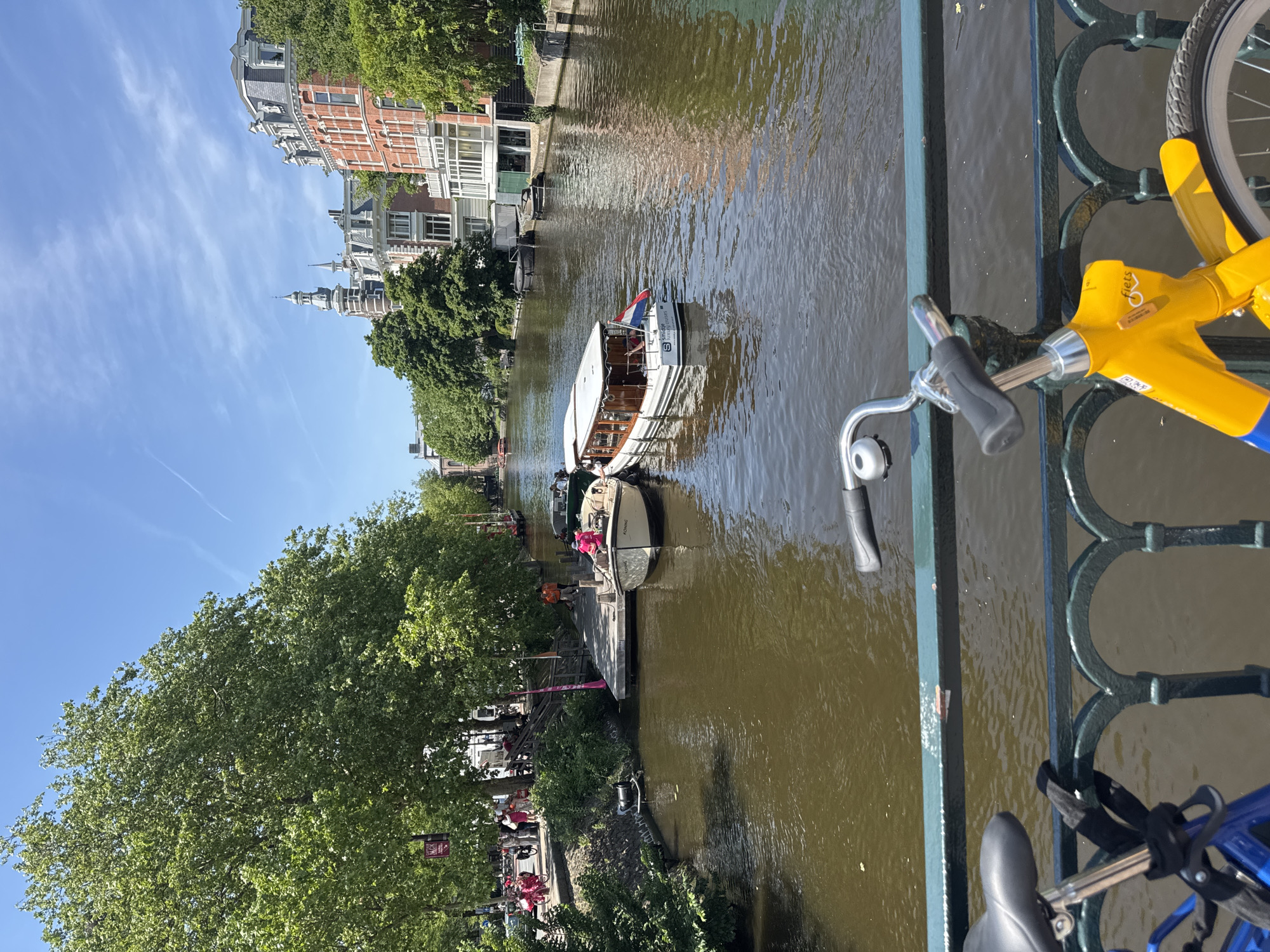 The image shows a canal scene in Amsterdam, Netherlands, taken from a bridge. On the right, a portion of a bicycle is visible, indicating the viewpoint. A canal flows beneath, with boats present. One boat is moving along the canal and another is docked at a platform along the side. People can be seen along the canal bank under the trees. Buildings line the other side of the canal, including buildings with peaked rooftops. The sky is blue.