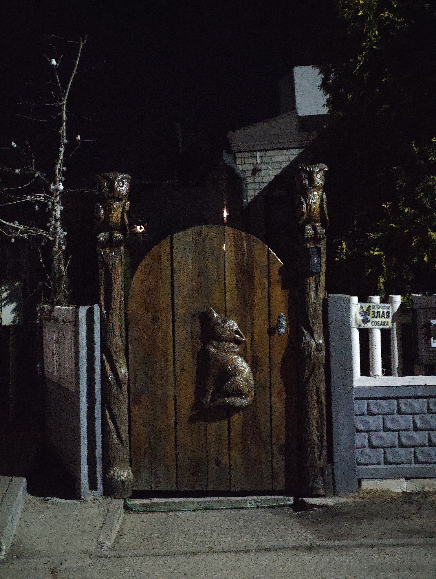 Captured at night in Парэчча, Belarus, the image depicts a rustic wooden gate flanked by carved owl figures atop wooden posts. The gate itself features a relief carving of a howling wolf. To the right, a sign warns of a "злая собака" (evil dog). A brick wall and a fence with white vertical slats surround the gate. Dim light illuminates the scene, revealing nearby trees and buildings in the background.