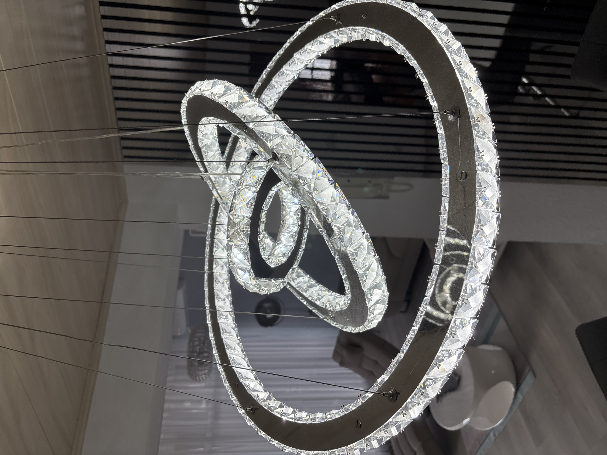 A modern crystal chandelier with three interlocking rings hangs from the ceiling in a well-lit room. The chandelier is adorned with numerous faceted crystals that reflect light, creating a dazzling effect. The rings appear to be made of a dark, metallic material. In the background, a light-colored ceiling and a wall with vertical dark stripes are visible. The room also contains furniture, including a sofa and a white table with a rounded design, suggesting it is a living or dining area. The overall ambiance is one of contemporary elegance.